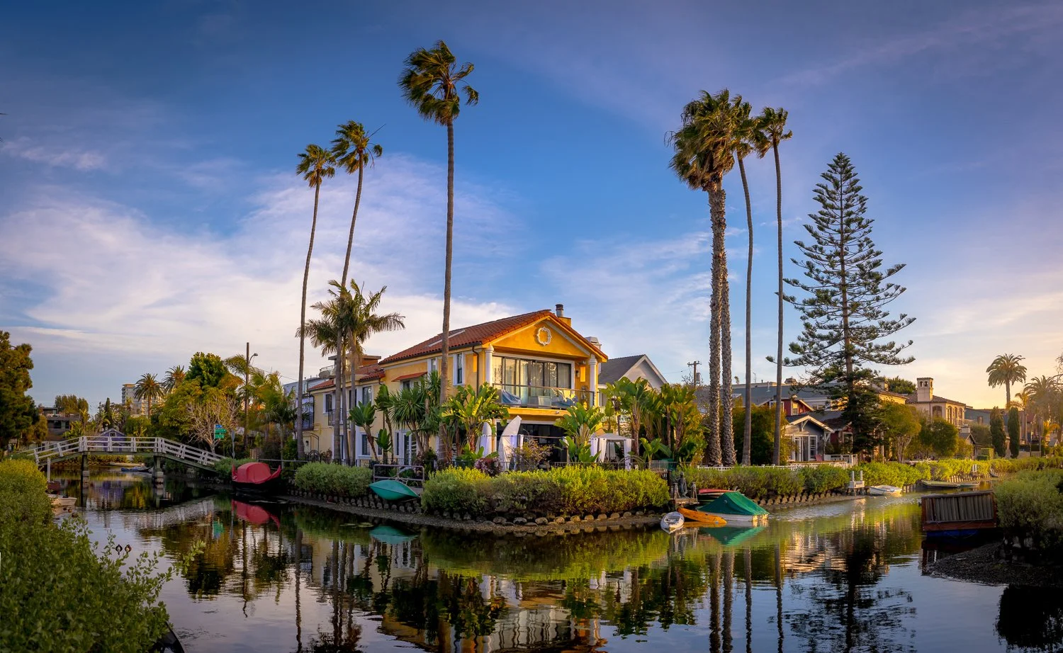 A scenic view of a house by a water canal with palm trees, boats, and lush greenery during sunset.