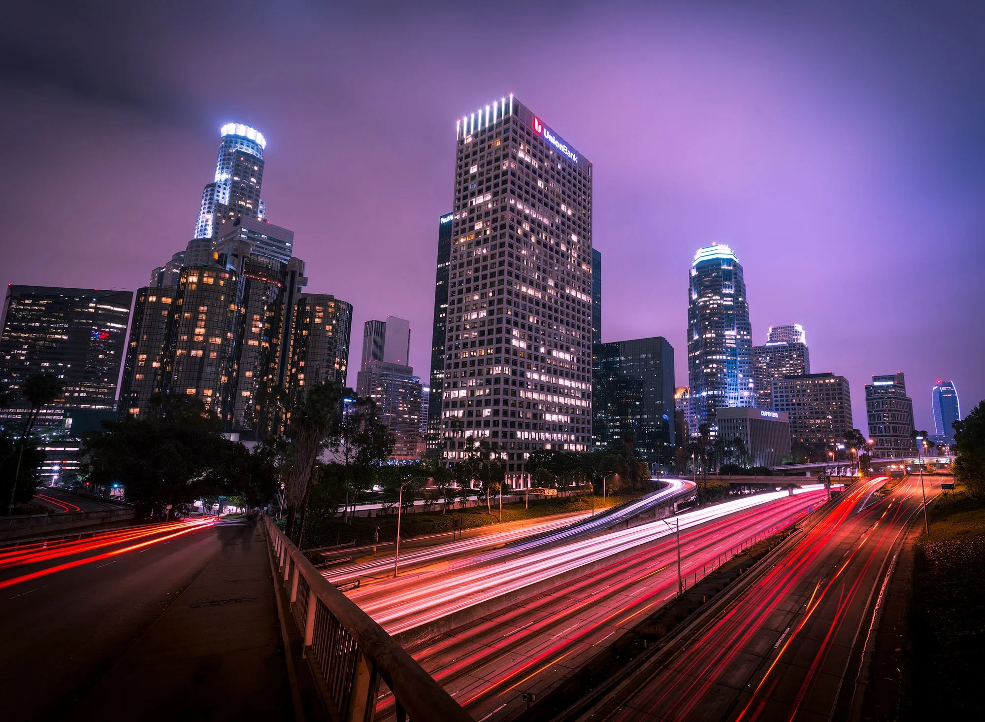 Nighttime cityscape of Los Angeles with illuminated skyscrapers and light trails from moving vehicles on highways.