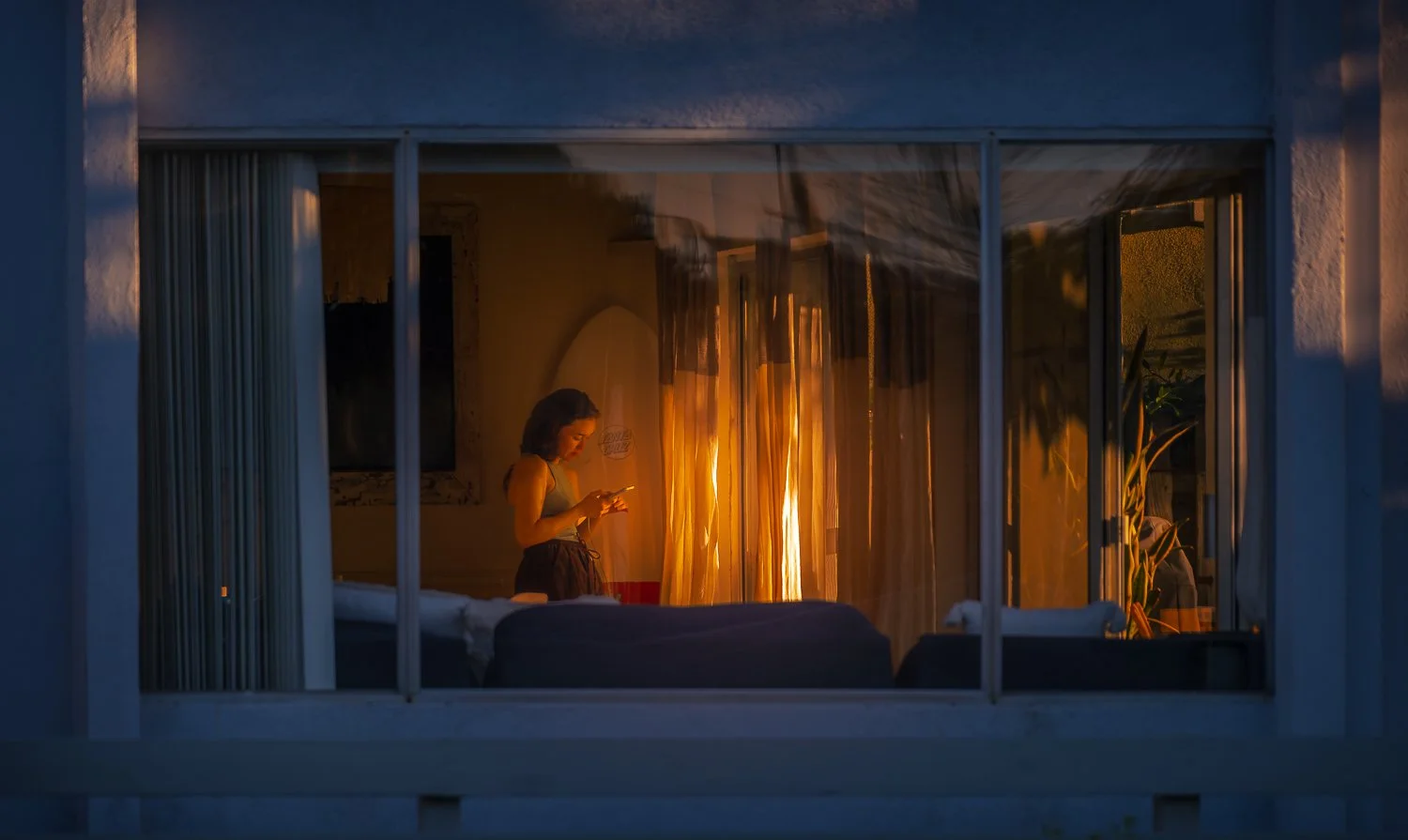 A person standing inside a room, looking at a phone, seen through a large window at dusk with warm lighting inside.