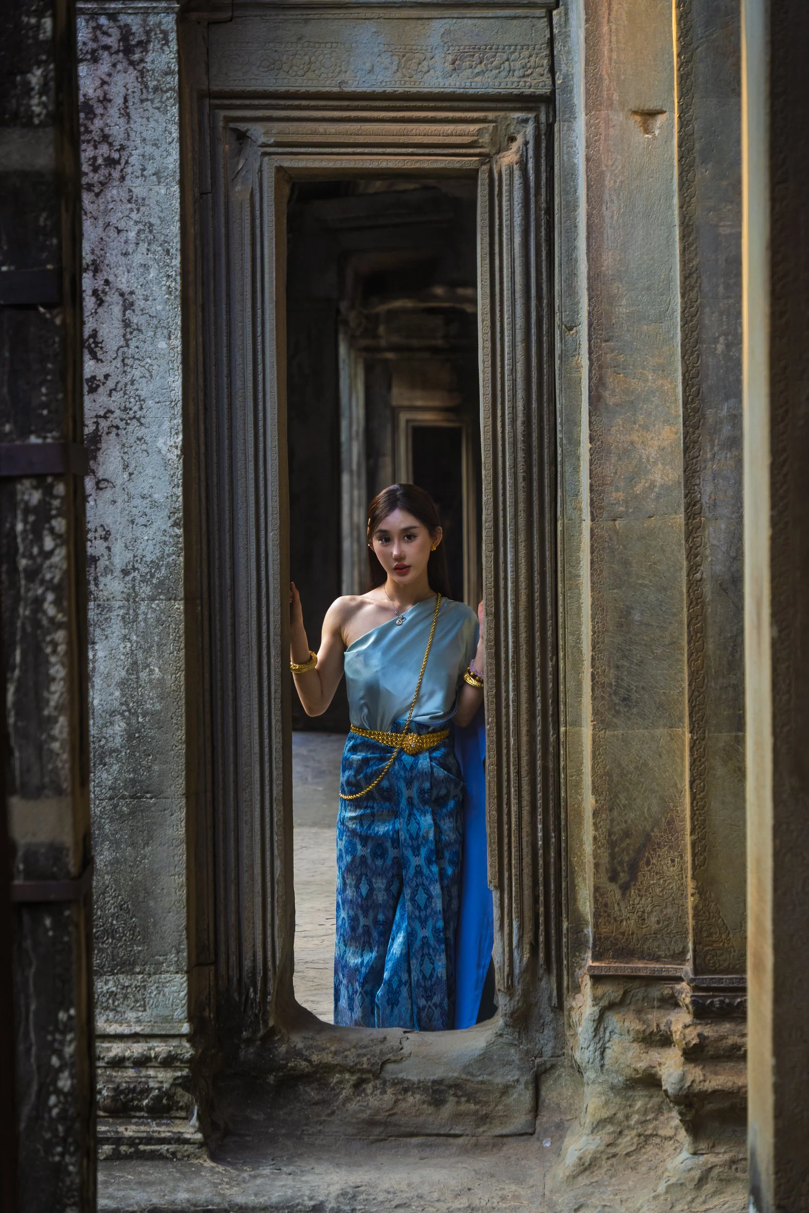 A young woman wearing traditional Thai attire stands in a narrow doorway surrounded by ancient stone ruins.