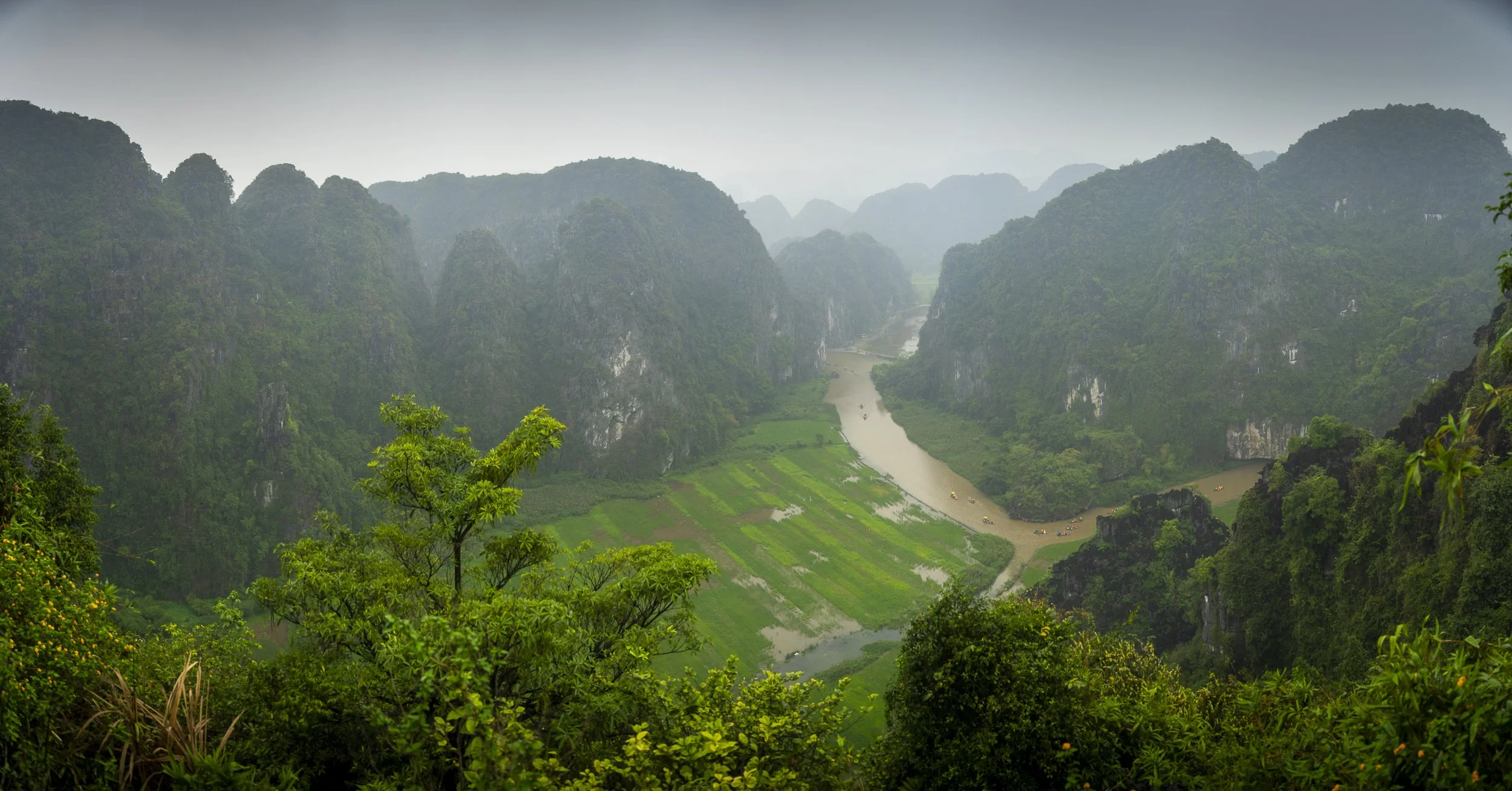 Lush green valley with a river winding through it, surrounded by tall, forested mountains under an overcast sky.