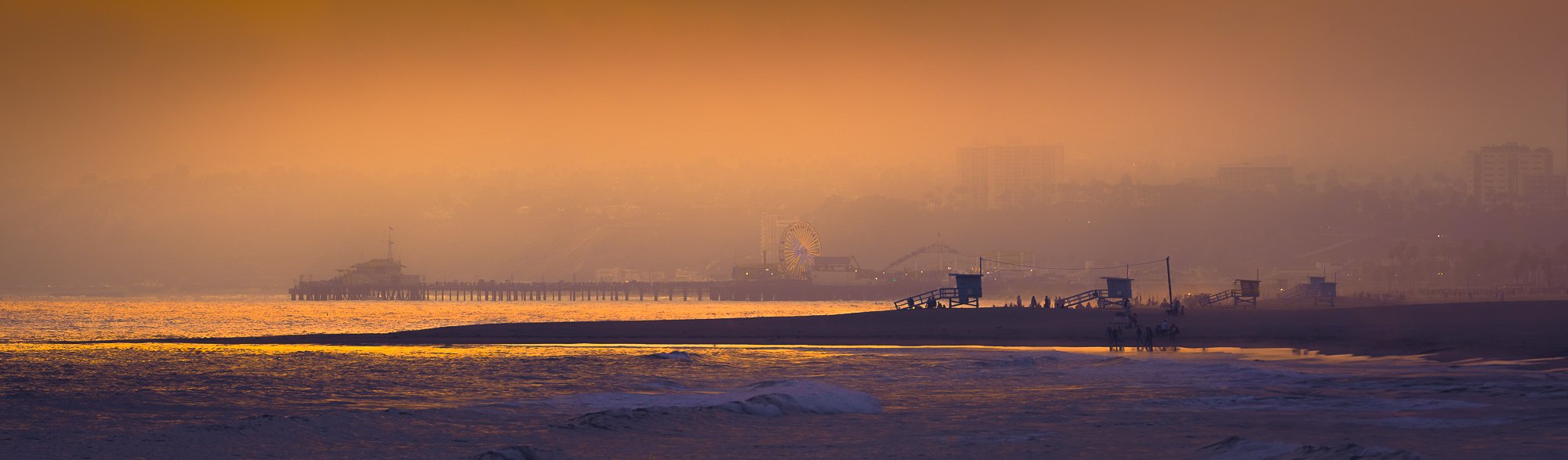 Sunset on a beach with a pier, amusement park rides, and lifeguard towers, with a hazy city skyline in the background.