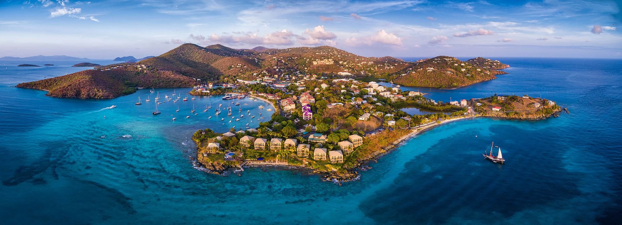Aerial view of a tropical island with lush green hills, white buildings, and a harbor filled with sailboats, surrounded by vibrant blue ocean waters.