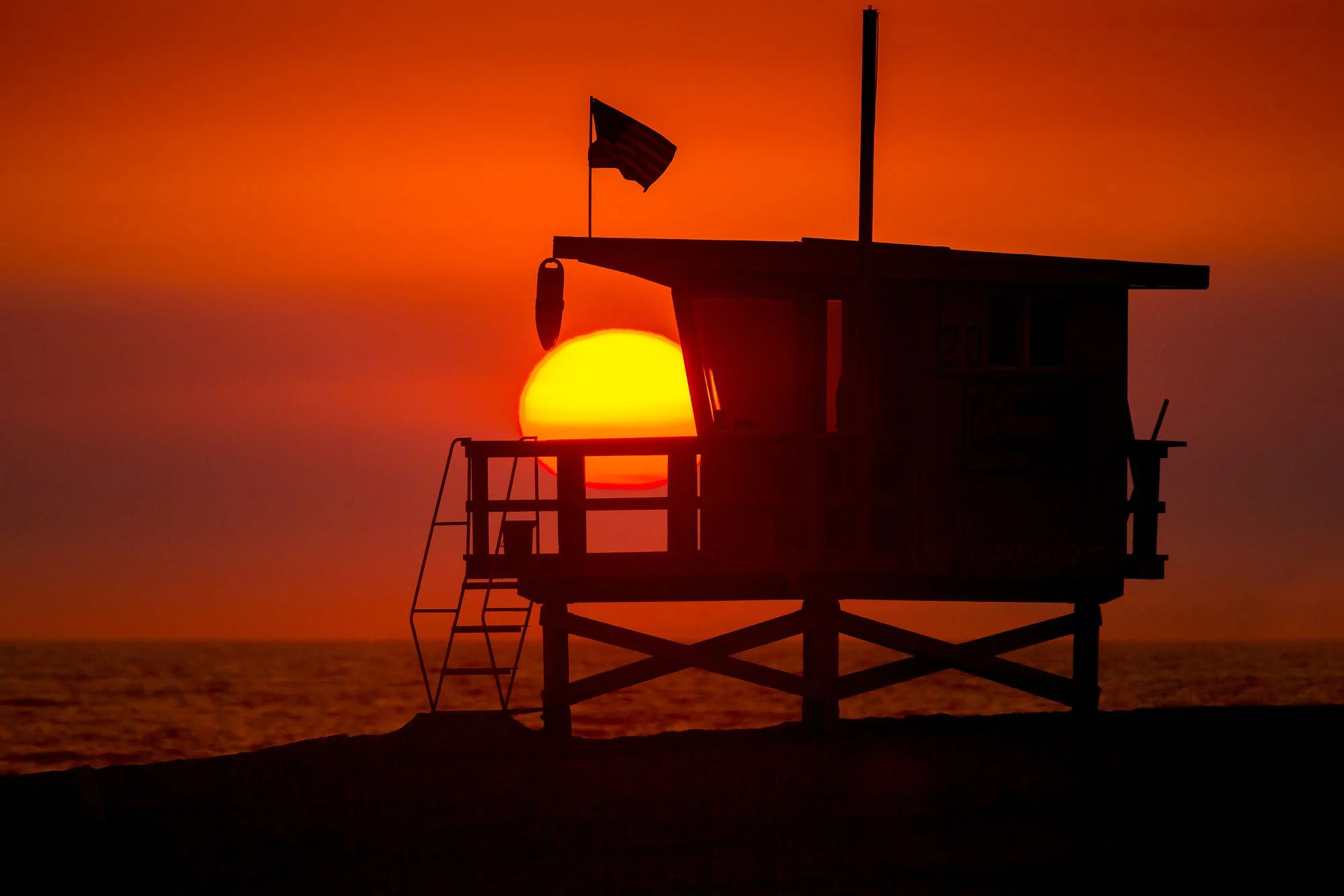 Silhouette of a lifeguard tower with a flag, against a bright orange sunset over the ocean.