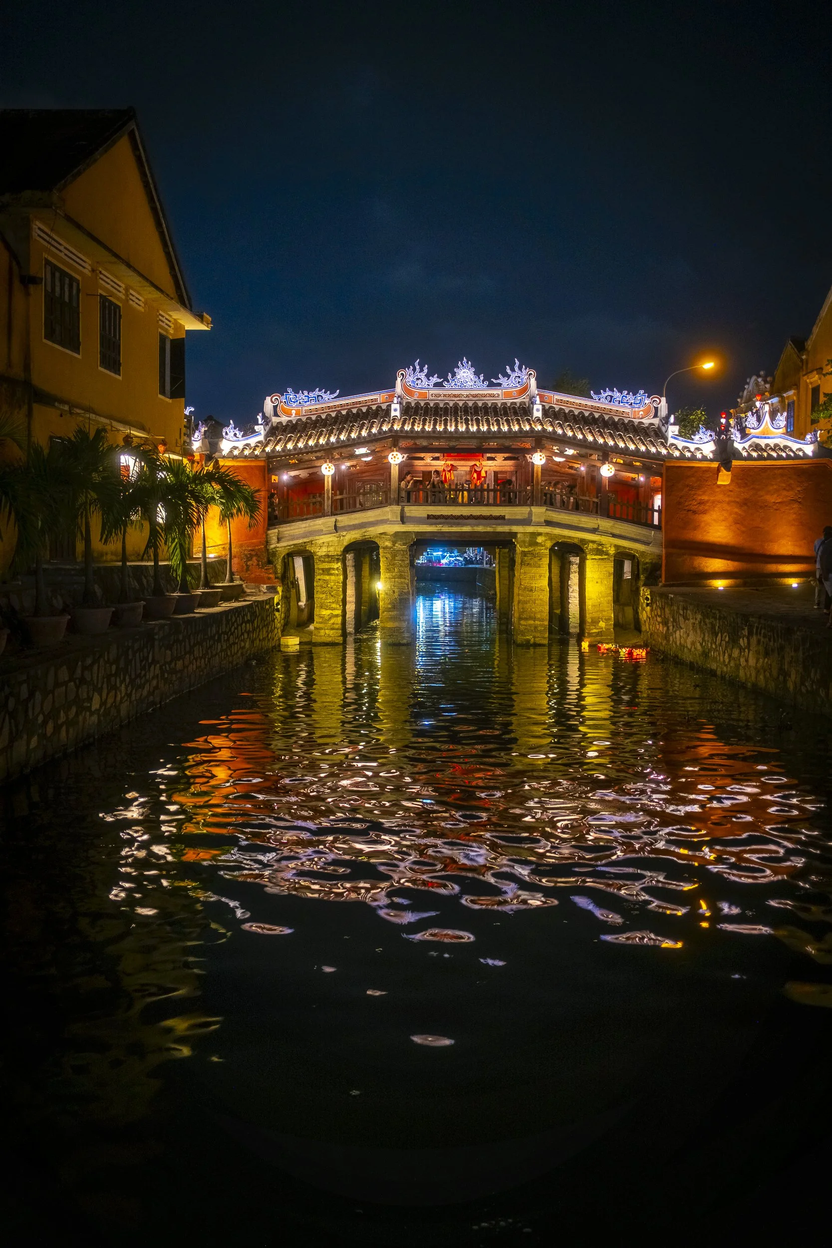 A night view of a traditional Chinese-style bridge with illuminated decorations, reflecting in the water below, surrounded by buildings and potted plants.