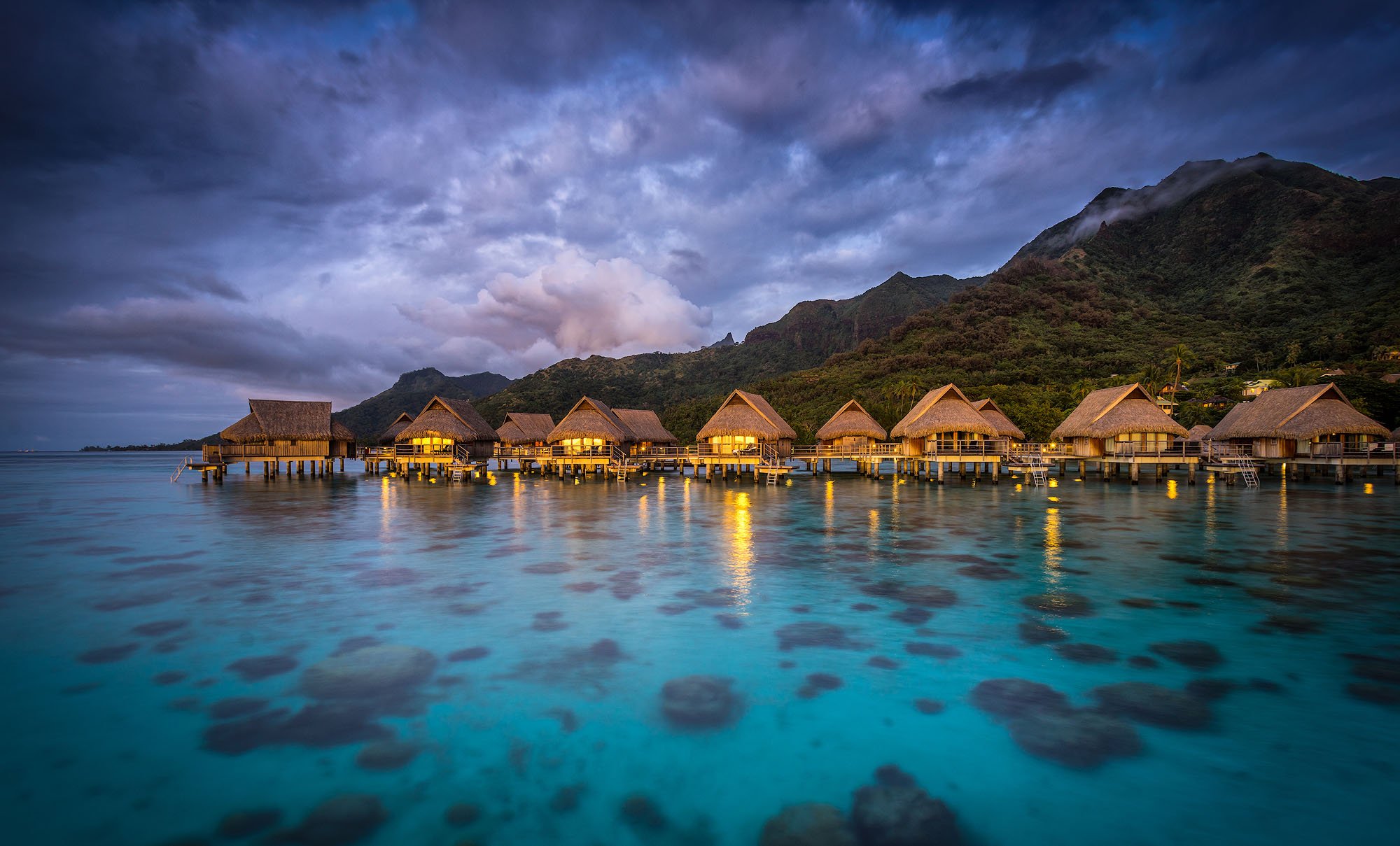 Overwater bungalows at dusk in a tropical location, mountains in the background and calm water reflecting the warm lights from the huts.