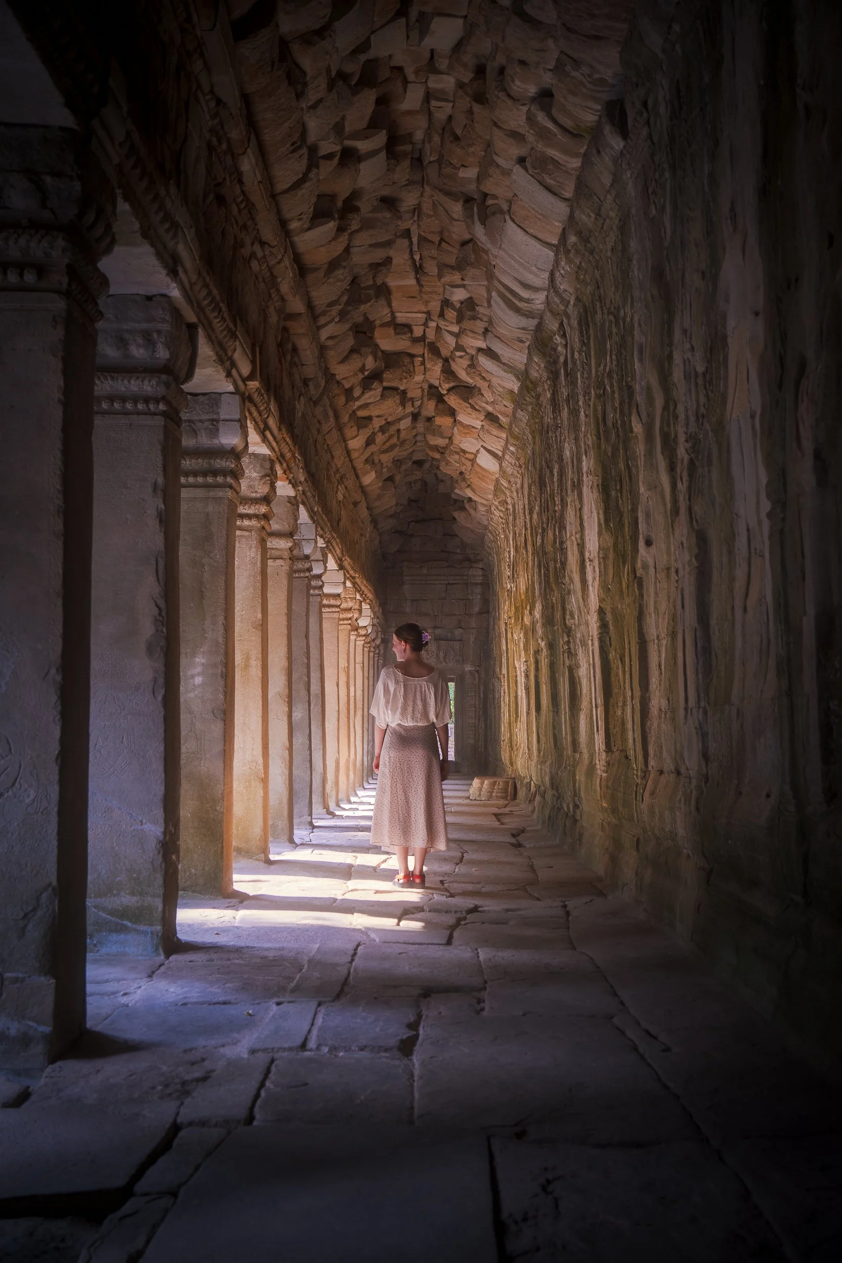 A woman in a white blouse and long skirt walking through an ancient stone corridor with columns on the left and mossy walls on the right, illuminated by soft natural light.