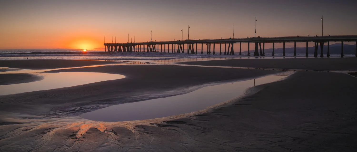 Sunset over a beach with a pier extending into the ocean, with wet sand and tide pools in the foreground.