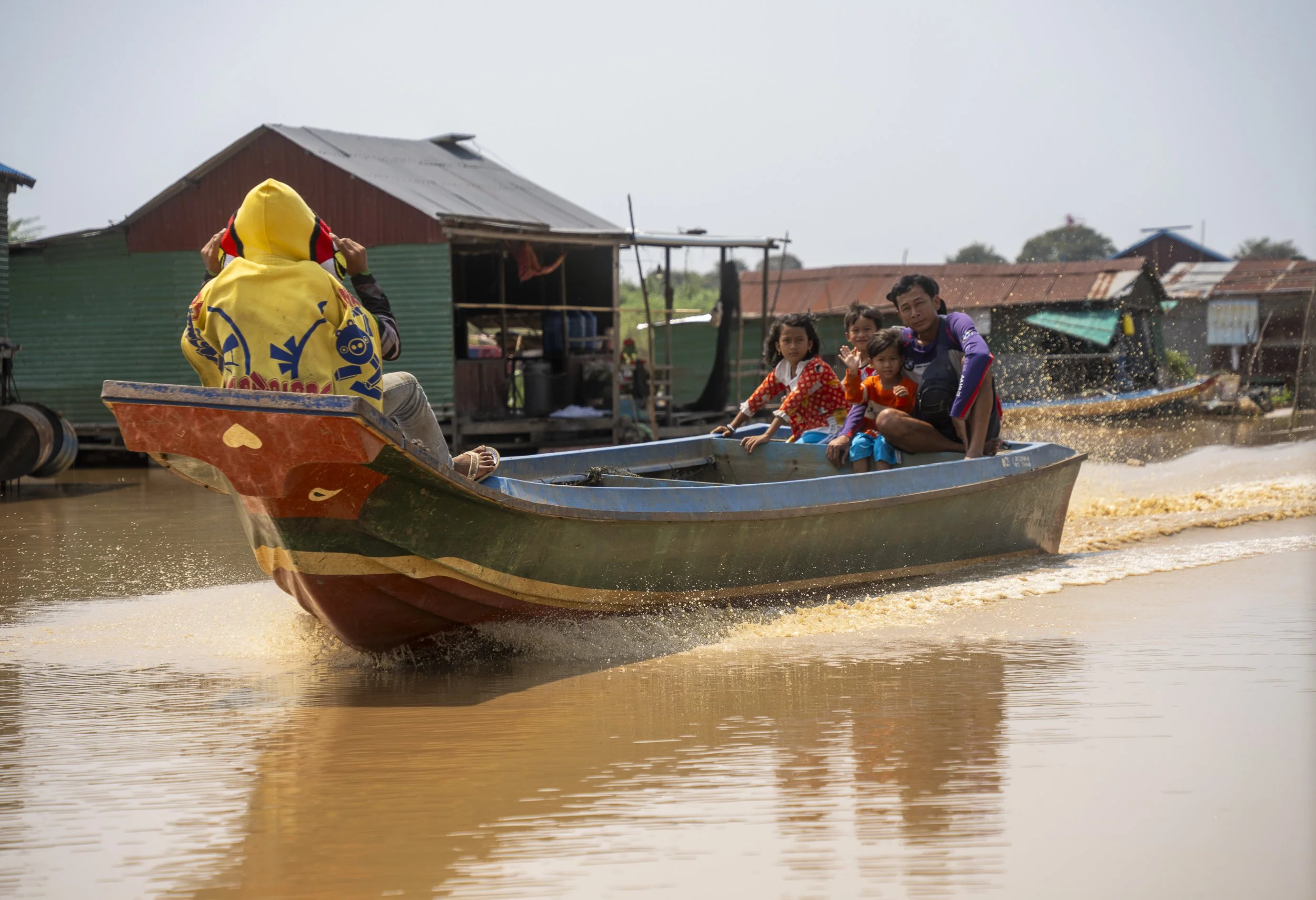 A family of five riding in a boat on muddy water, with houses on stilts in the background; the father is steering, and the children are waving.