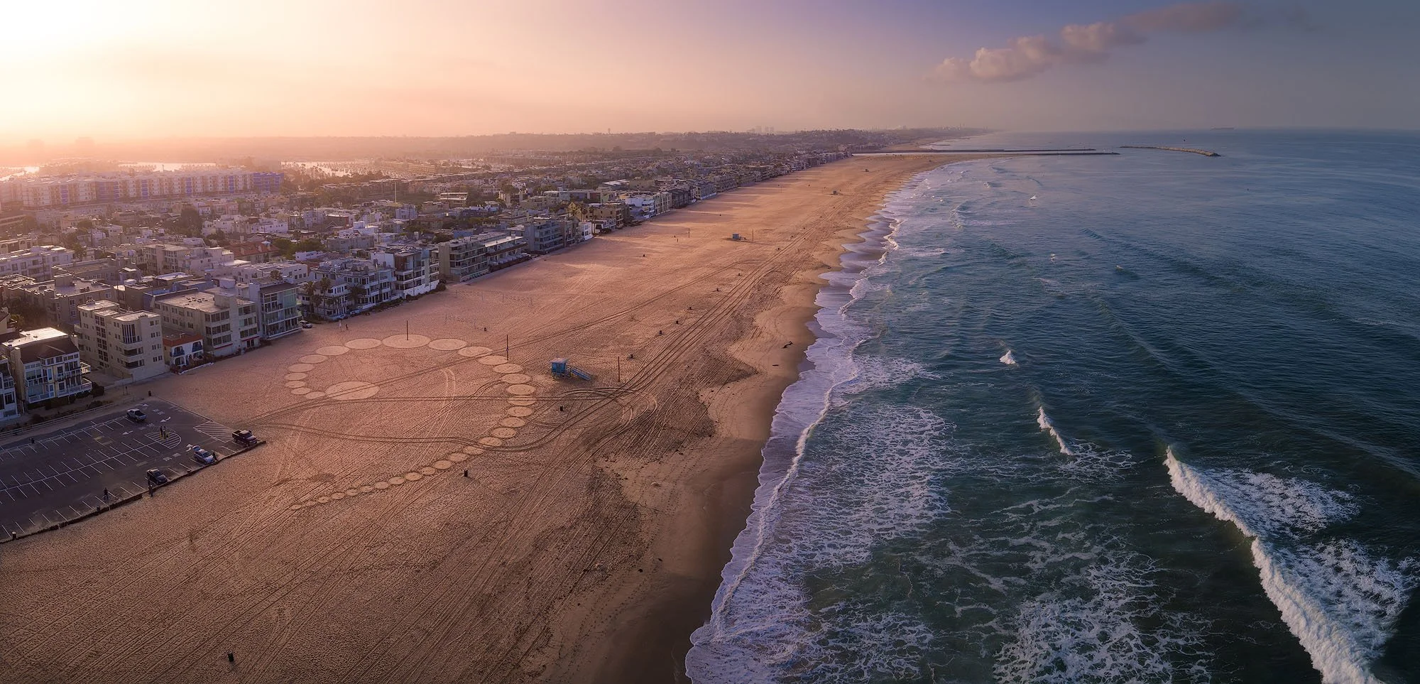 Aerial view of a sandy beach with a city behind and ocean waves in the foreground, taken at sunset.