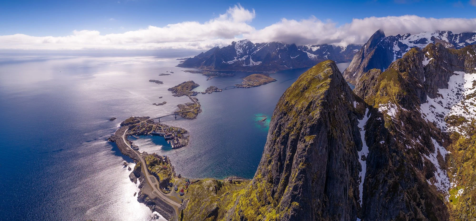Aerial view of a rugged, mountainous coastline with snow-capped peaks, a deep blue fjord, and a small village with a bridge connecting islands.