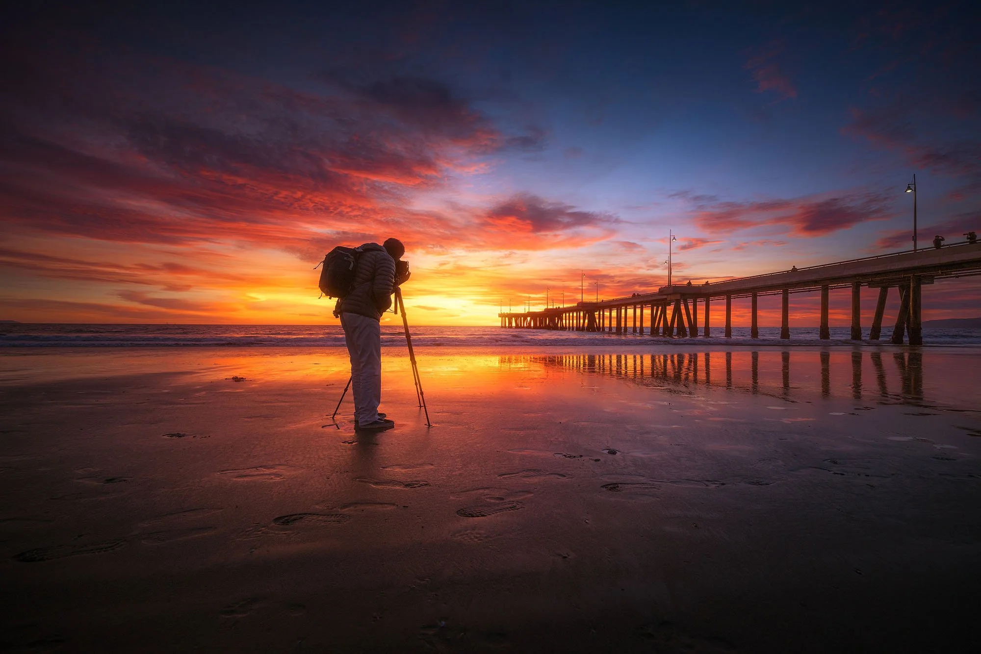 Person taking photos on the beach at sunset near a pier.
