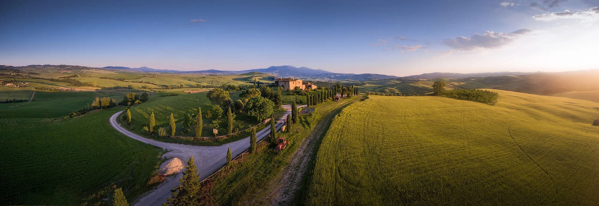 A scenic landscape of rolling green fields, a winding dirt road, and a large house with cypress trees in the countryside, with mountains in the background, during sunset.