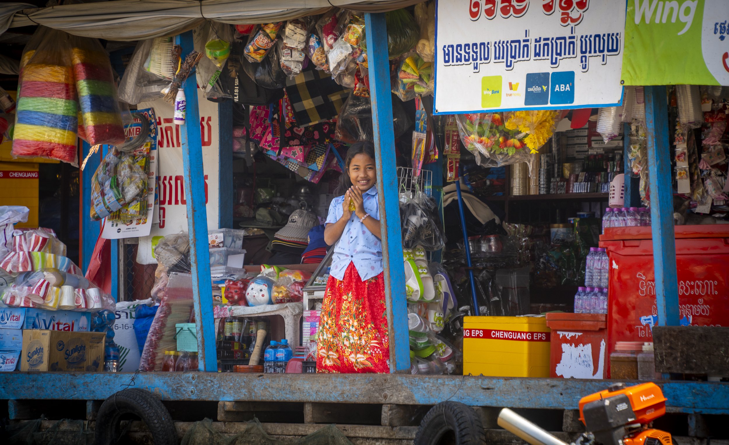 A young girl smiling and holding her hands together standing inside a small colorful shop with various goods, including toys, bags, and household items, in a market setting.