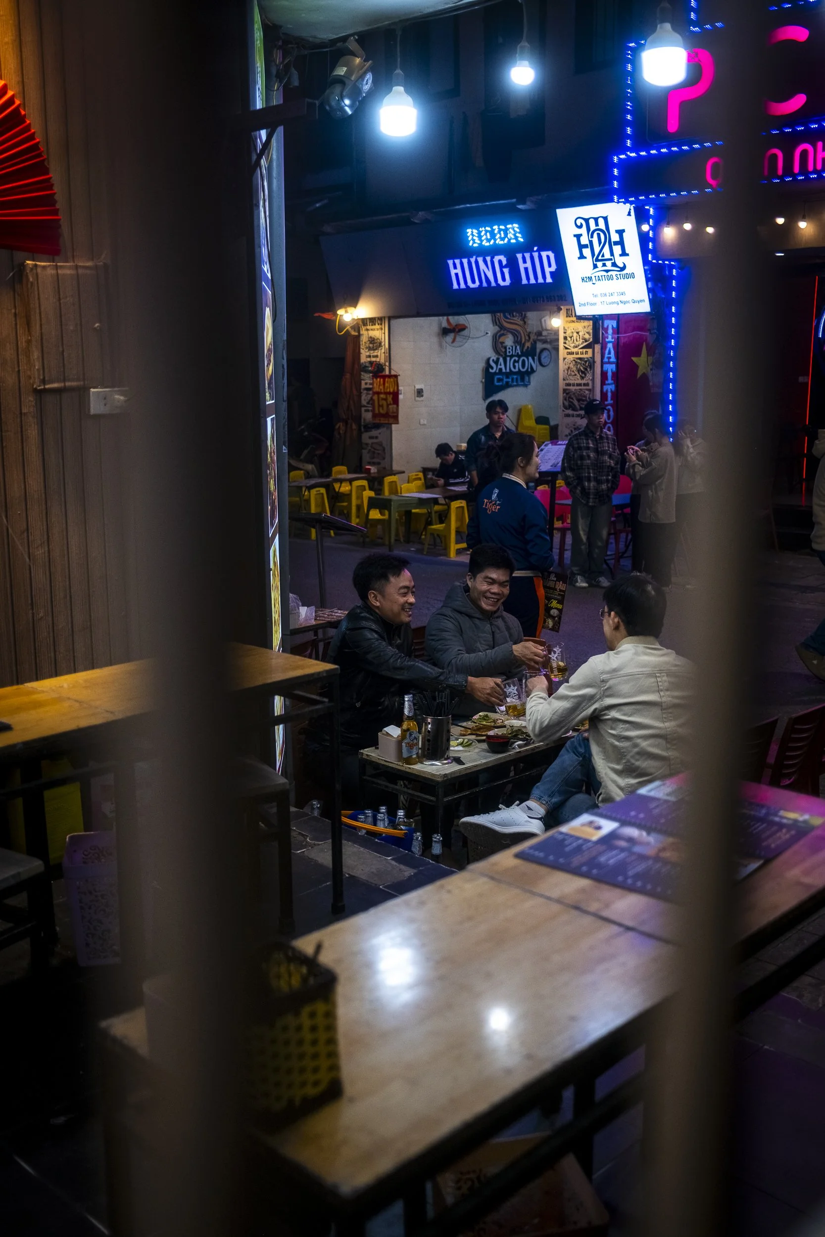 A group of four men sitting at a table in a restaurant, laughing and clinking glasses, with a busy street scene with neon signs visible outside.