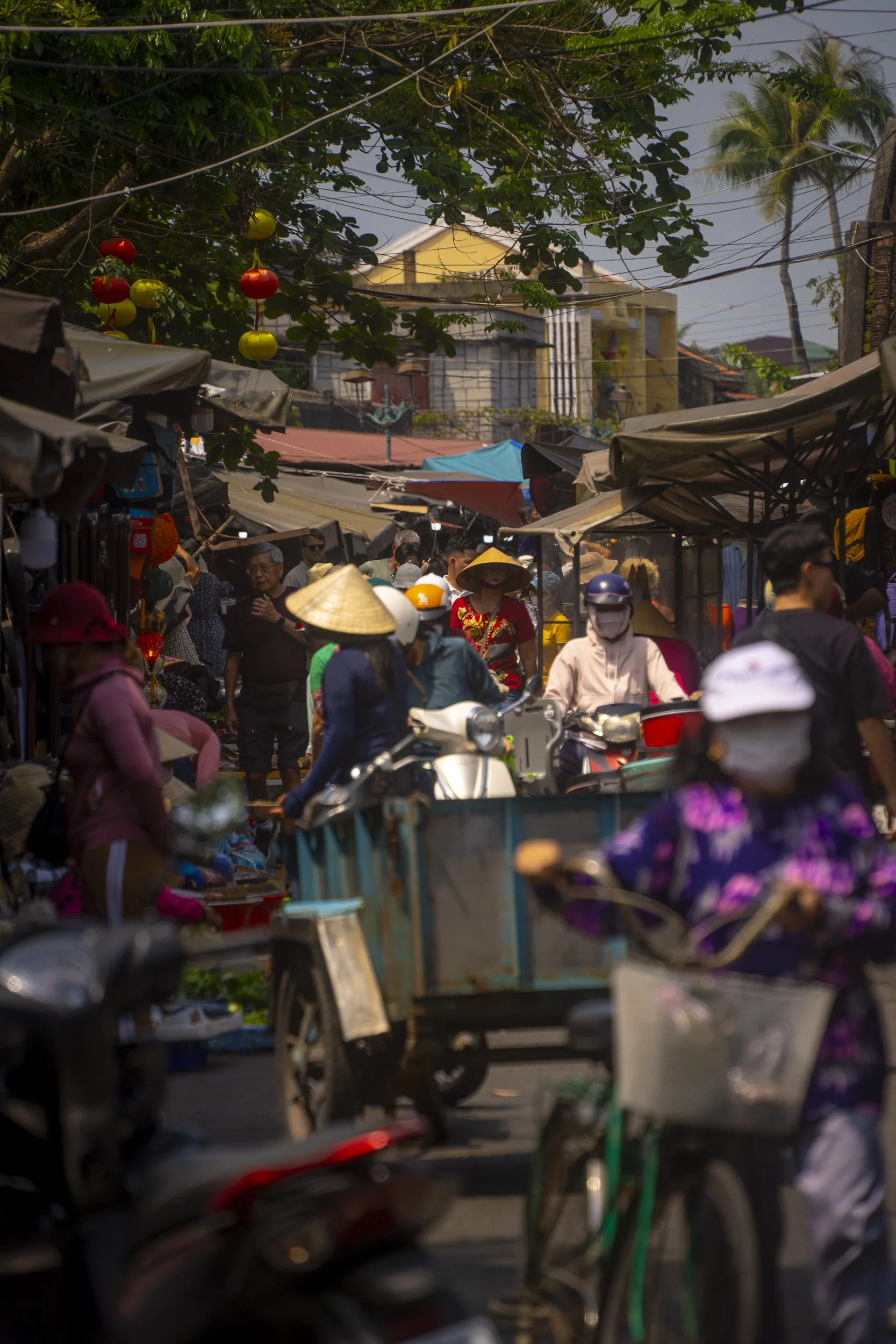 A busy market street filled with people, motorbikes, and carts. Some people wear traditional conical hats, and colorful lanterns hang from trees. Market stalls line both sides of the street under awnings.