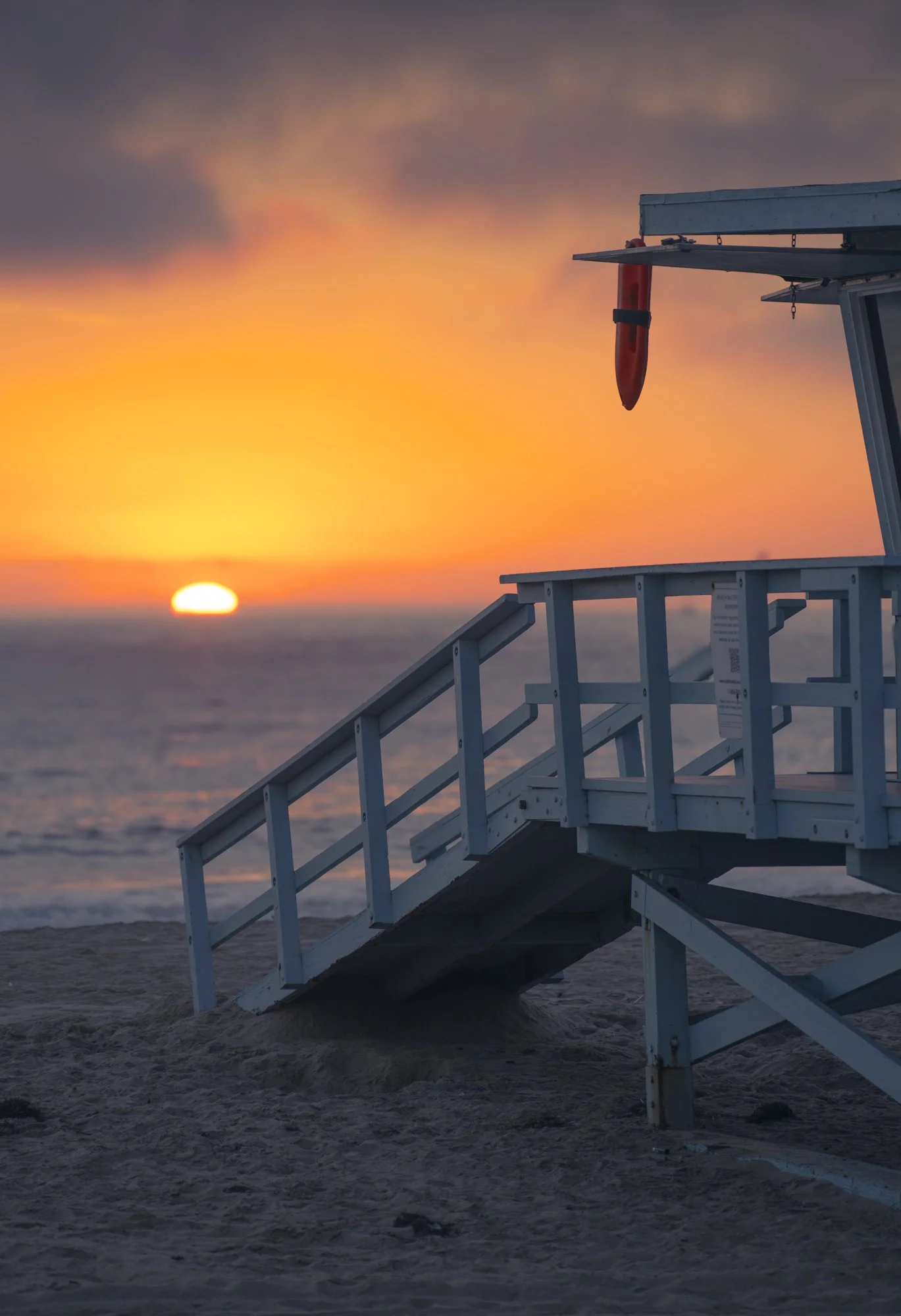 Empty lifeguard chair on the beach at sunset with the sun near the horizon, cast in warm orange and yellow hues.