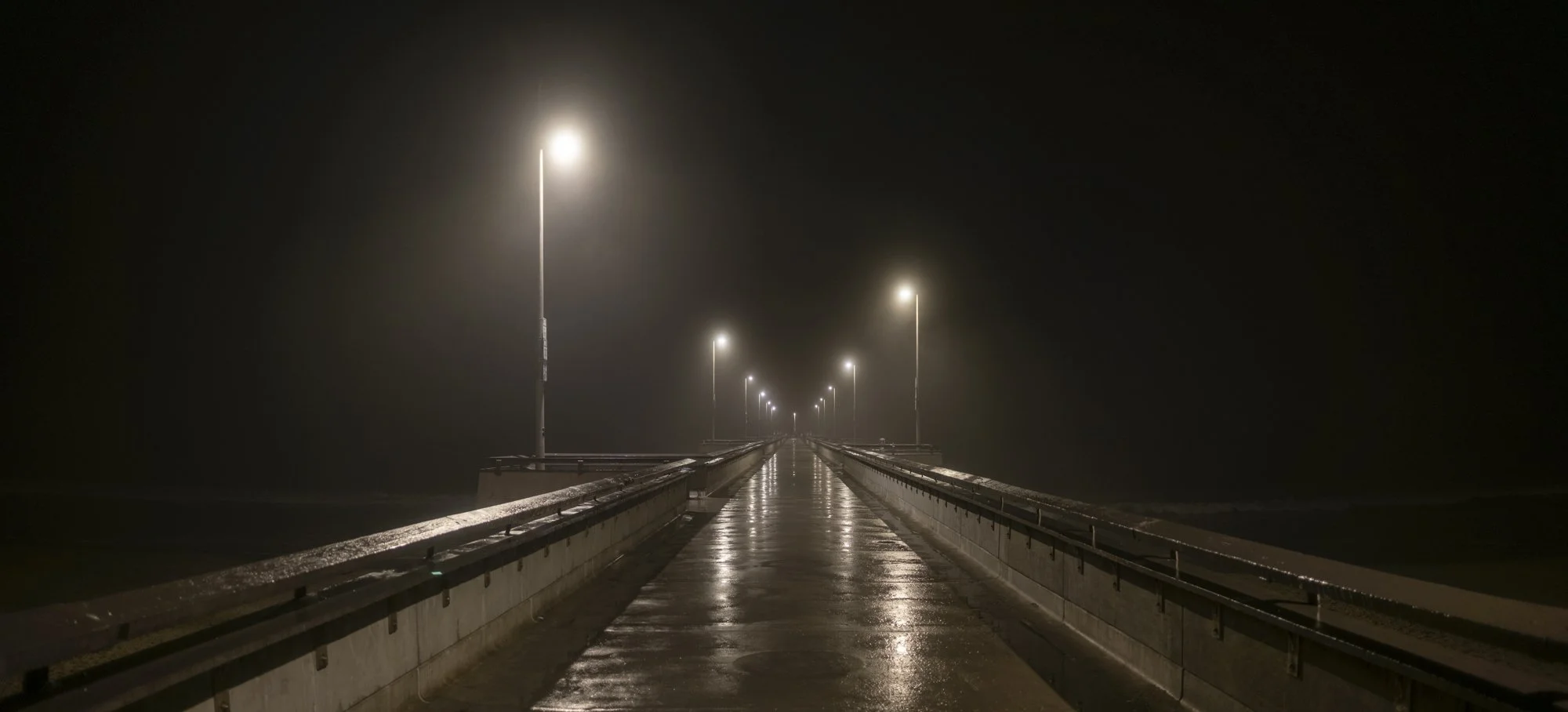 A foggy, empty pier with wet surface and streetlights extending into the distance, creating a moody nighttime scene.