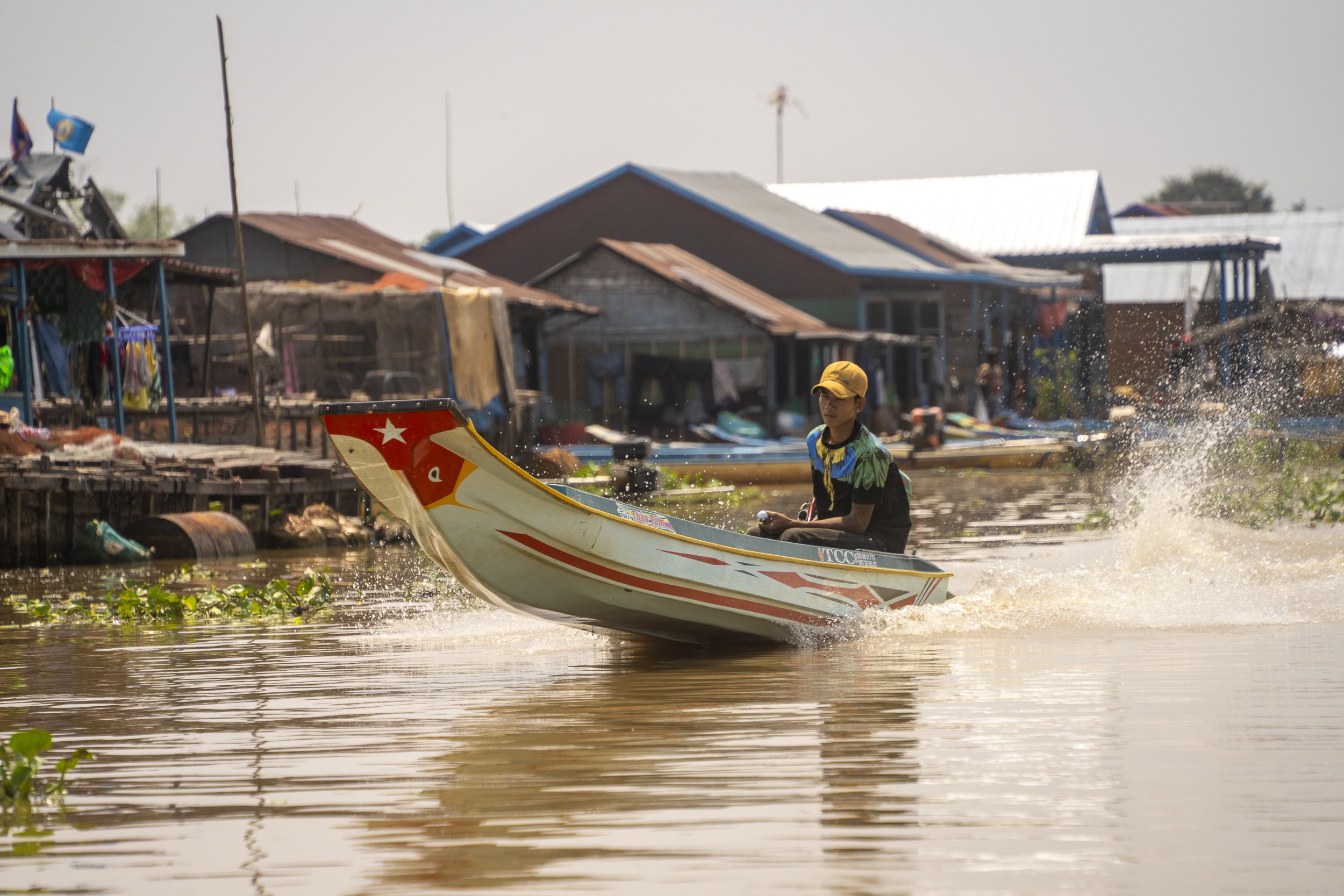 A young man riding a speedboat on a river in a rural area, with stilt houses and boats in the background.