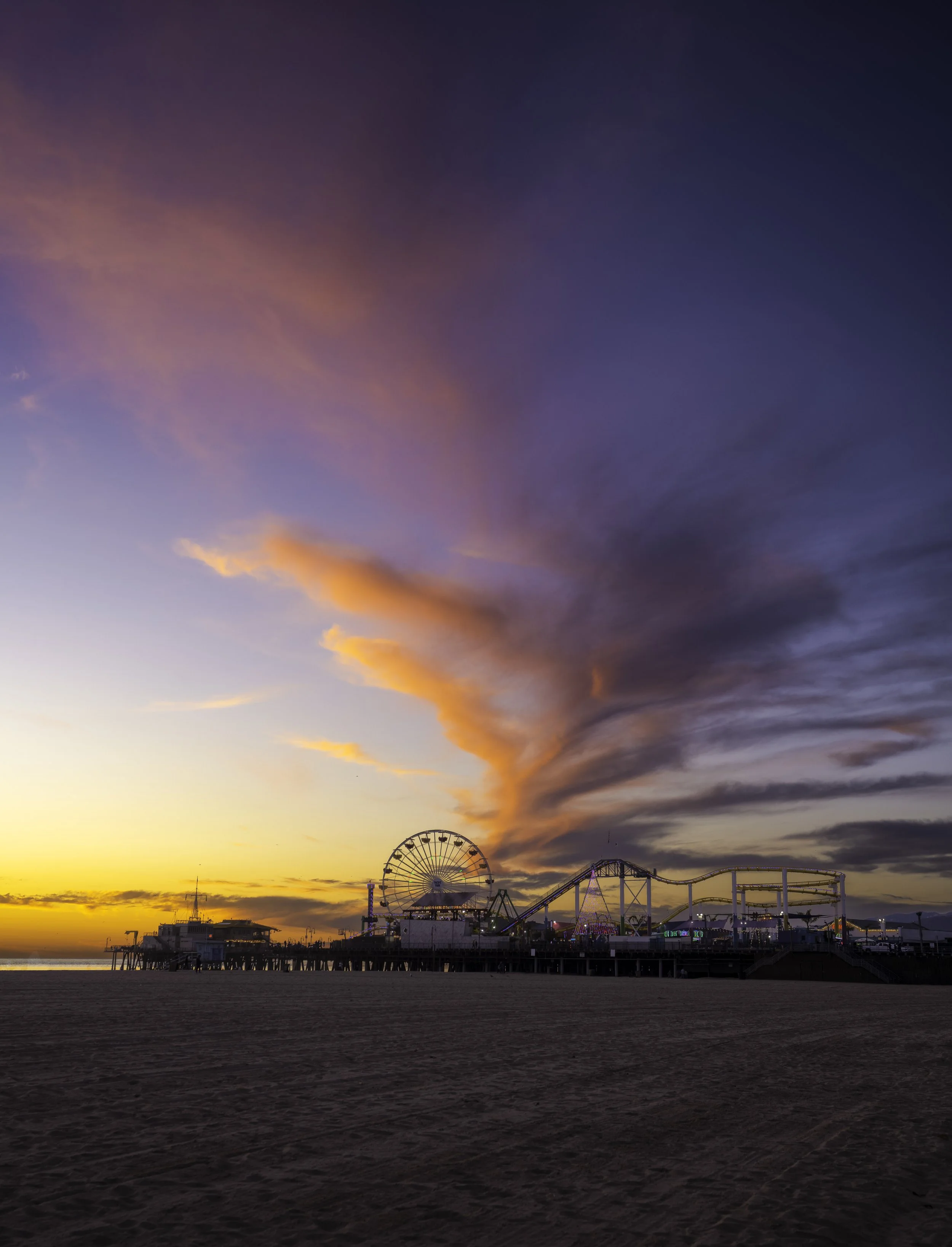 Sunset over an amusement park on the beach, with a Ferris wheel and rollercoaster rides visible, under a colorful sky with pink, purple, and orange clouds.