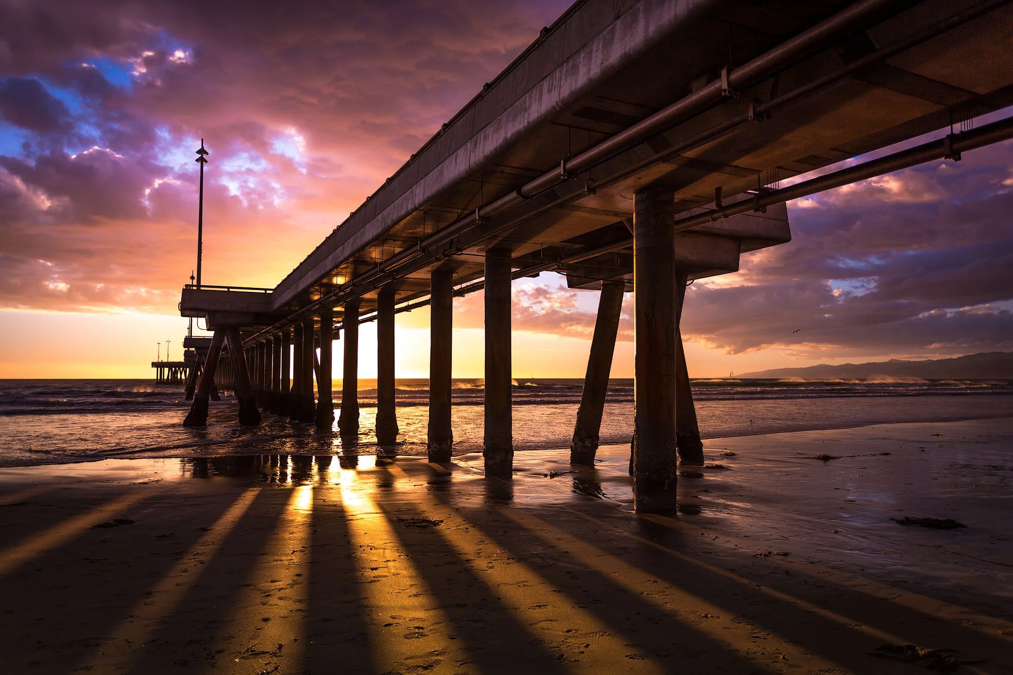 Sunset view of a pier extending into the ocean, with long shadows cast on the sandy beach and colorful clouds in the sky.
