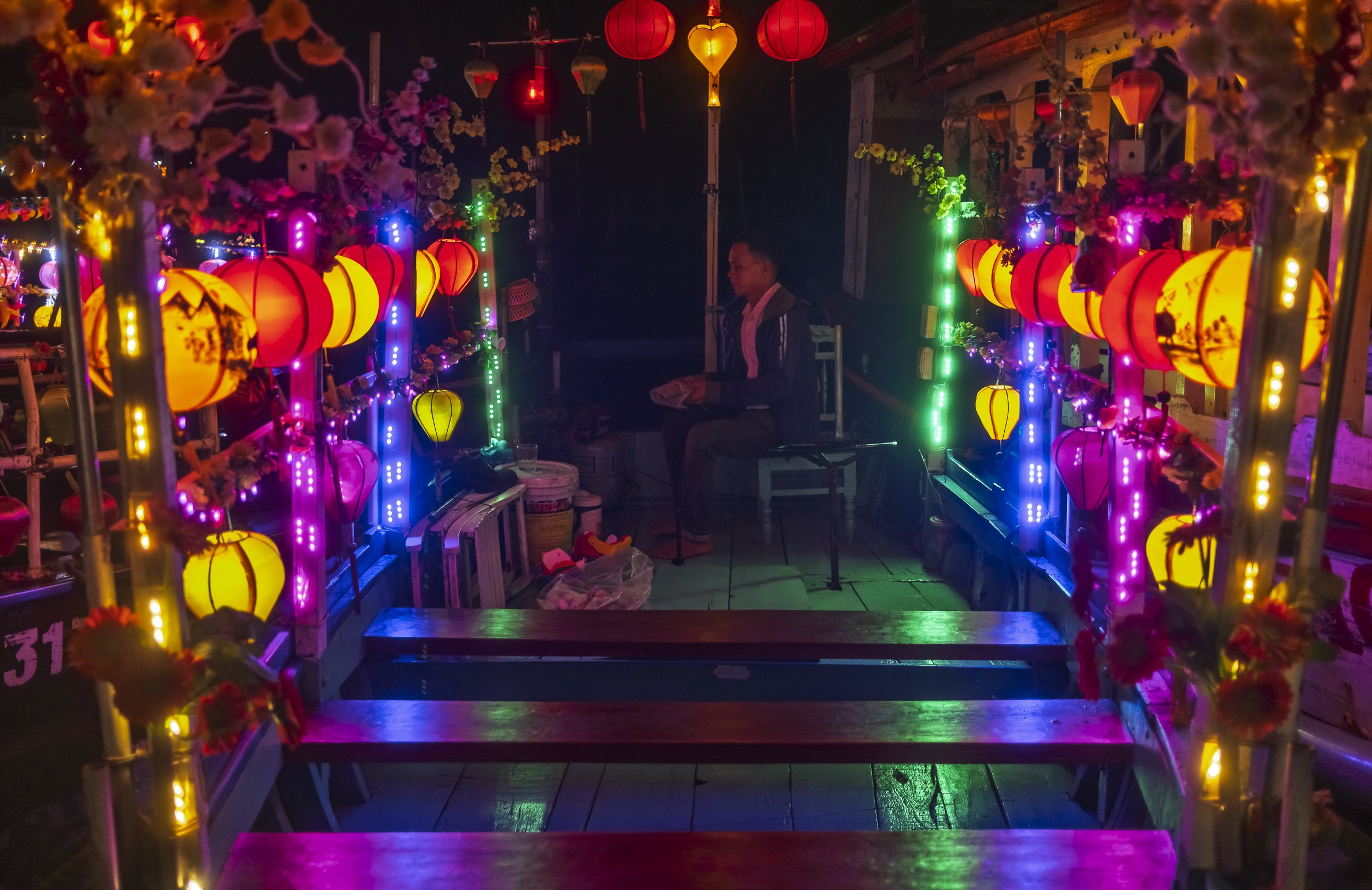A young person is sitting on a chair inside a colorful lantern-lit booth at night. The booth is decorated with illuminated lanterns in red, yellow, and purple, with floral accents and multicolored LED lights, creating a festive atmosphere.