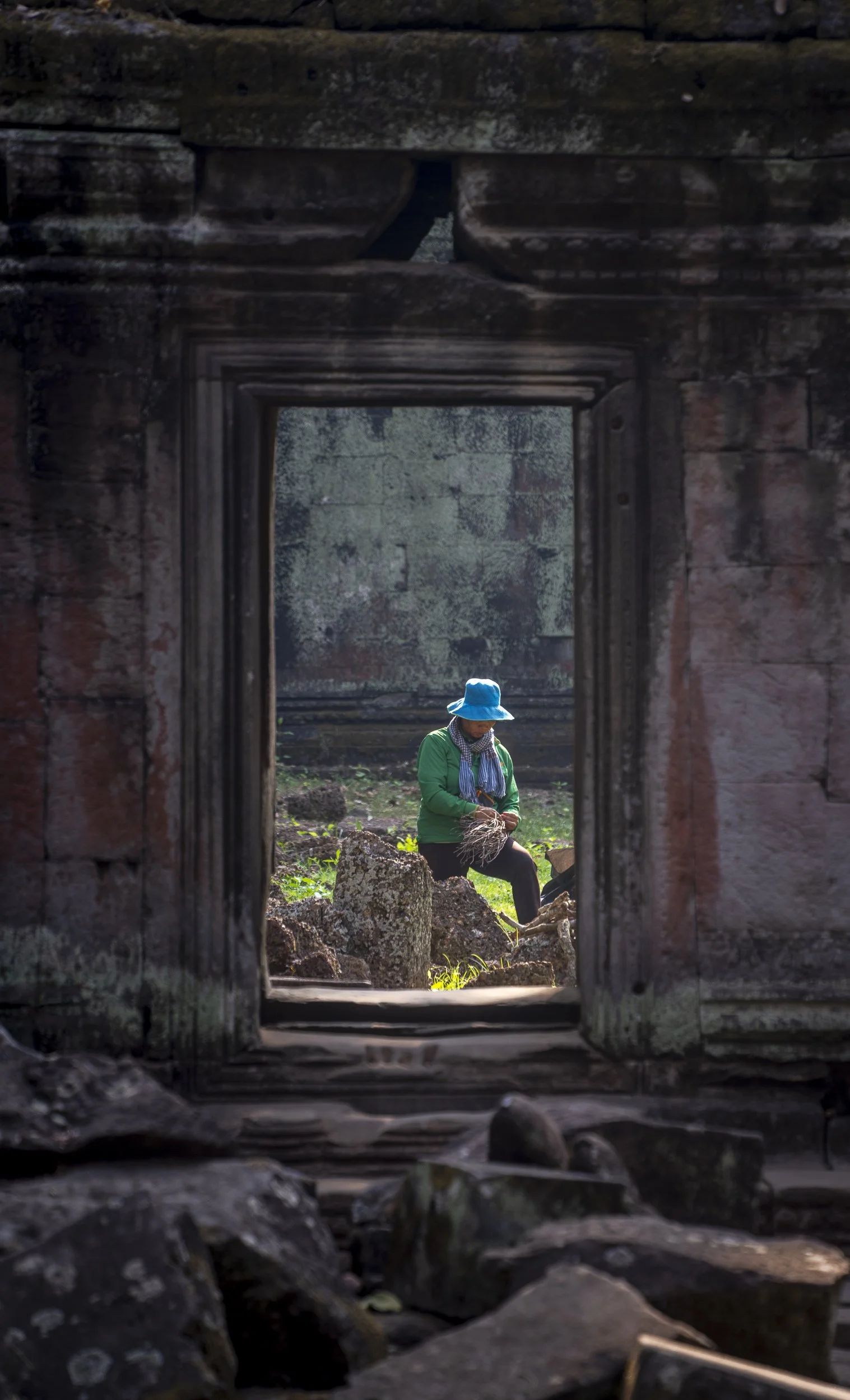 A person wearing a blue hat and green jacket is seen through an ancient stone window, working with a bundle of plant material outdoors.
