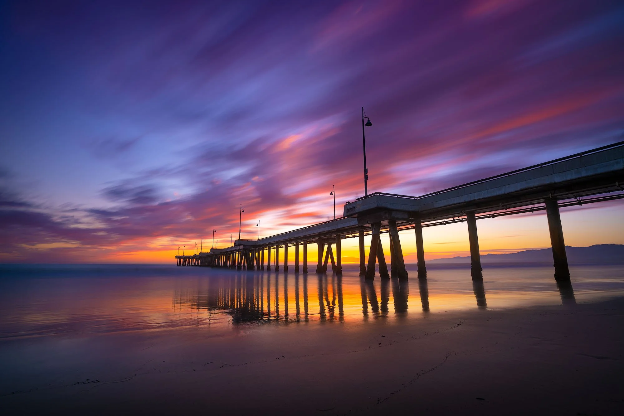 A pier extending into the ocean at sunset with colorful clouds in the sky and their reflection on the water.