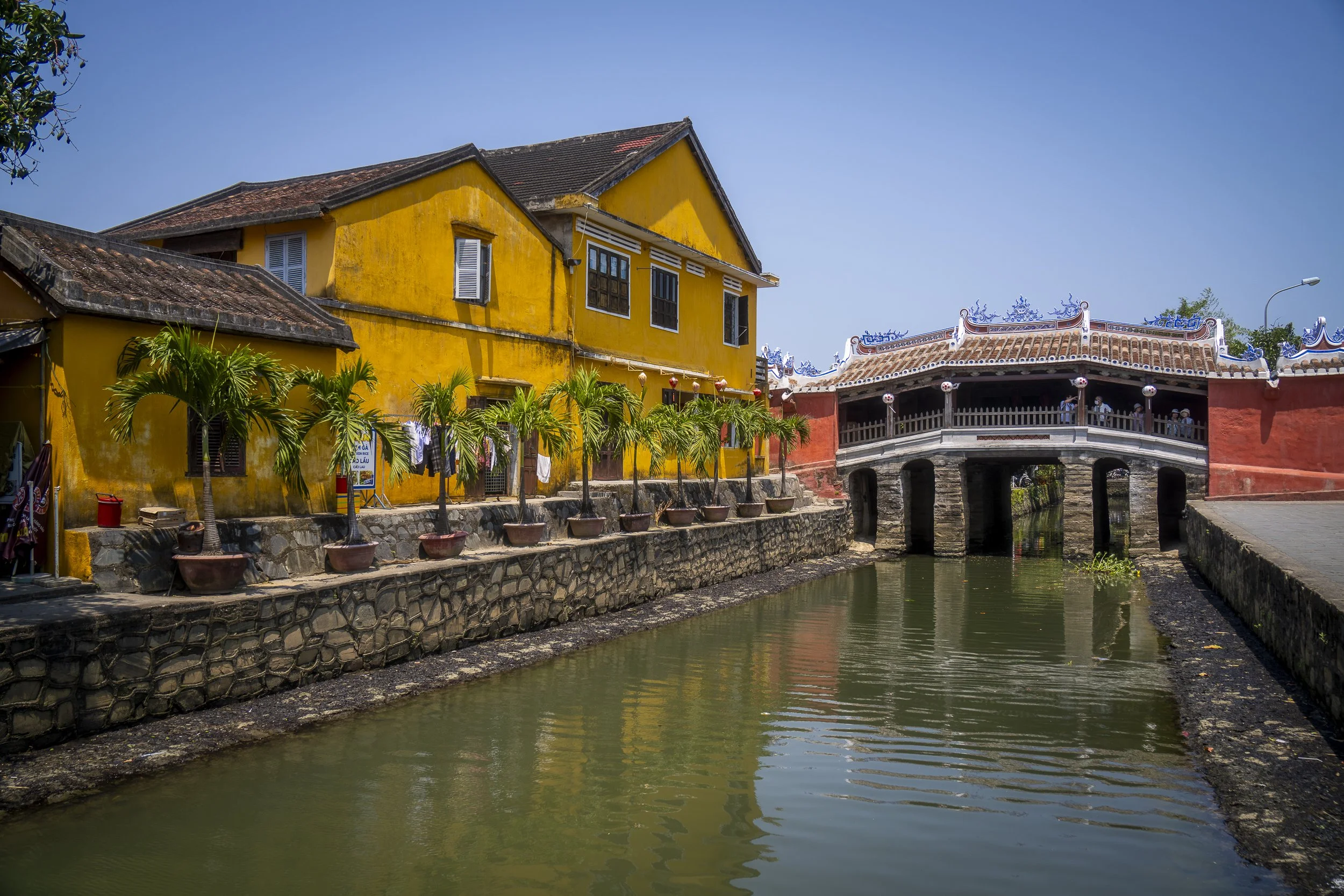 Colorful canal scene with a yellow building, potted palm trees, and a red bridge over water under a clear blue sky.