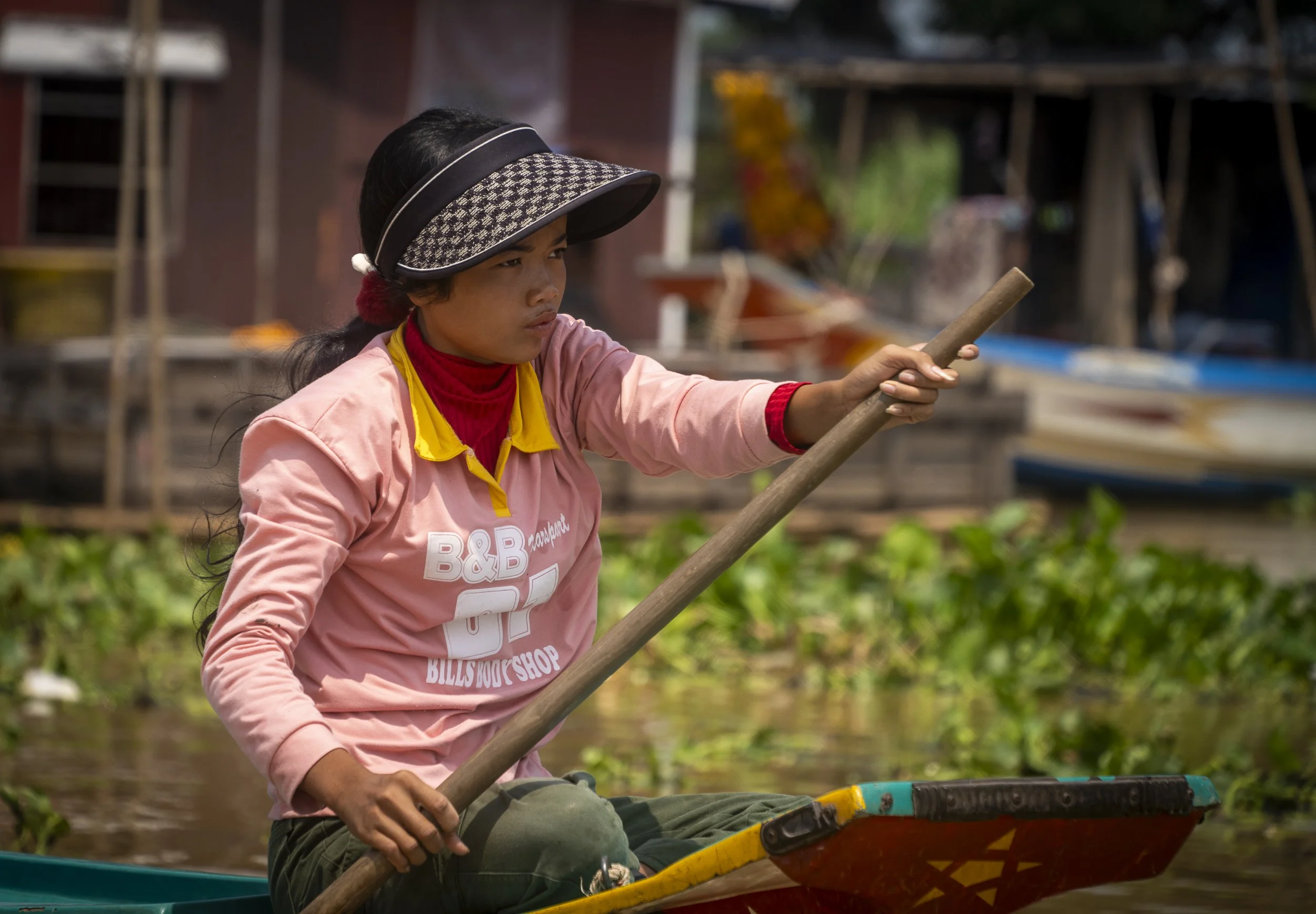 A woman paddling a small boat with a wooden oar in a rural area with houses and boats in the background.