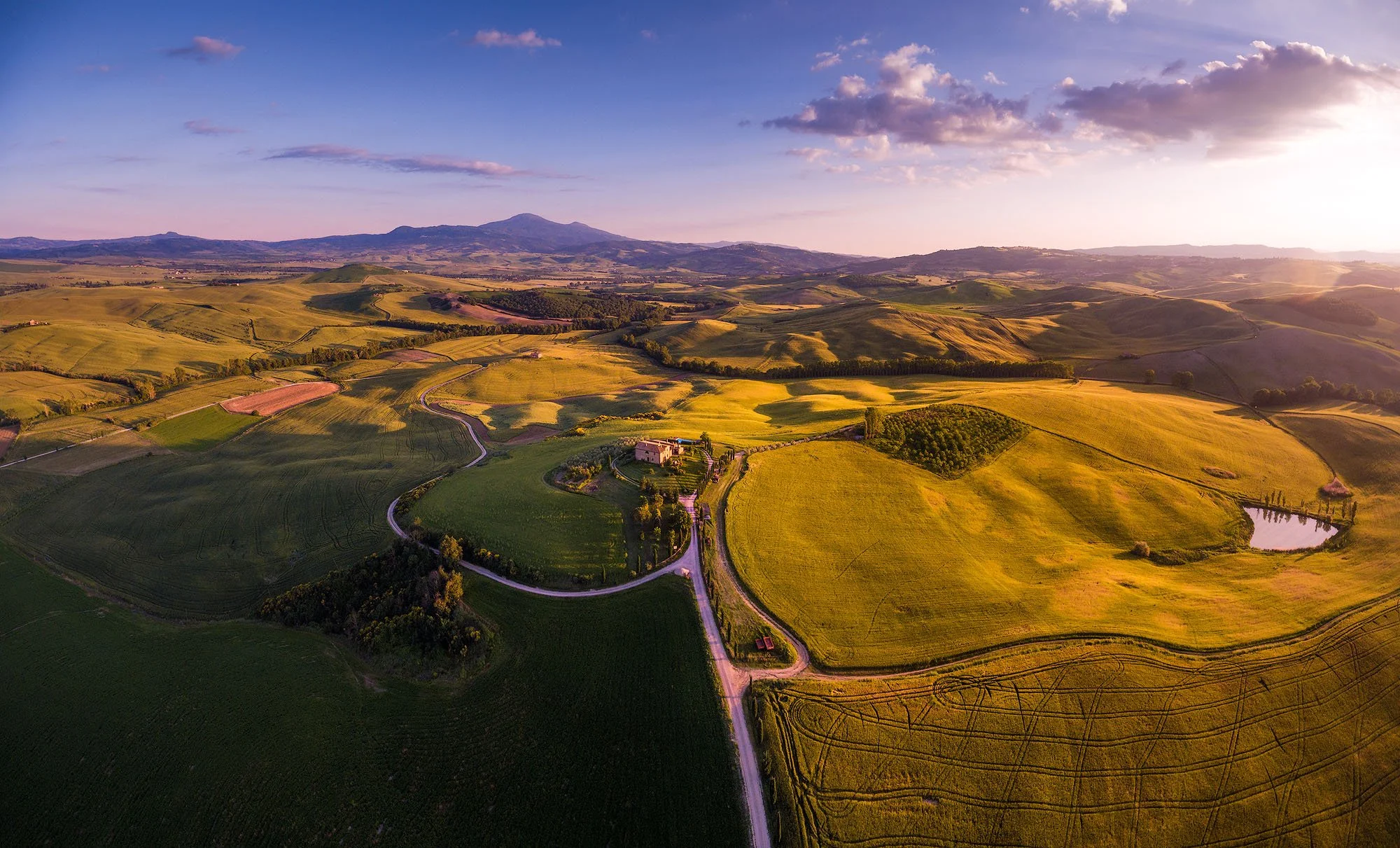 Aerial view of rolling green and yellow fields, winding roads, a pond, and distant mountains at sunset.