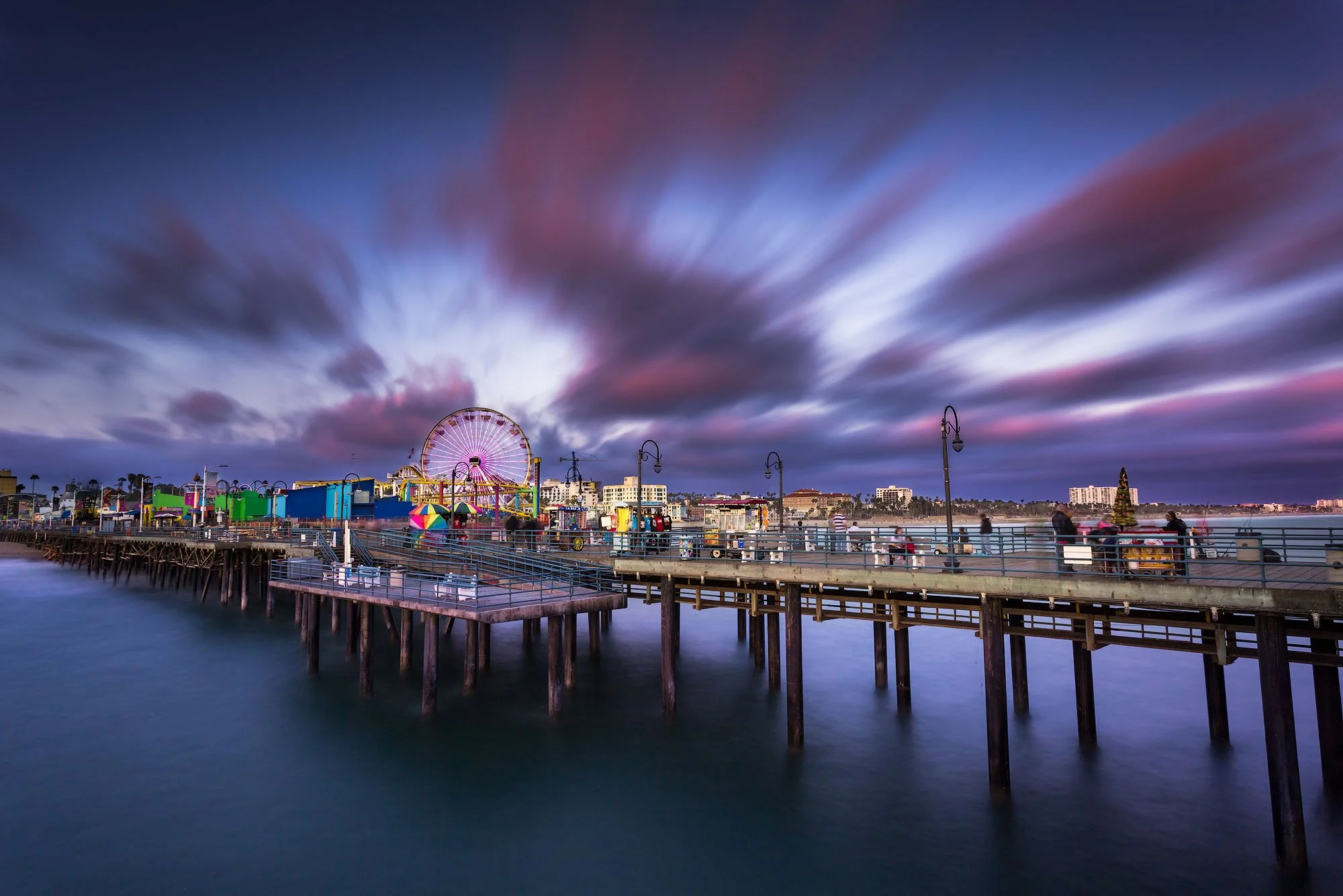 Long exposure photograph of an amusement park on a pier at dusk with colorful rides, including a Ferris wheel, under a cloudy sky with pink and purple hues.