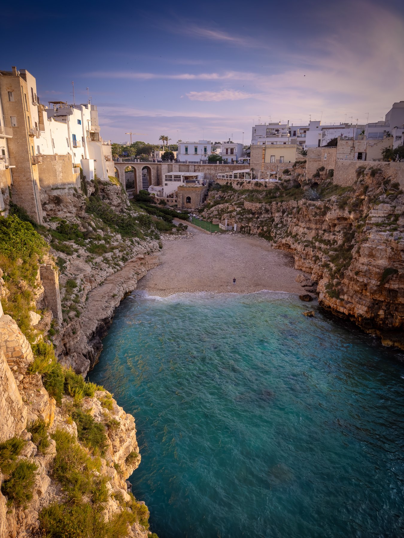 A rocky coastal cove with turquoise water, a small beach, and surrounding white buildings on cliffs under a partly cloudy sky.