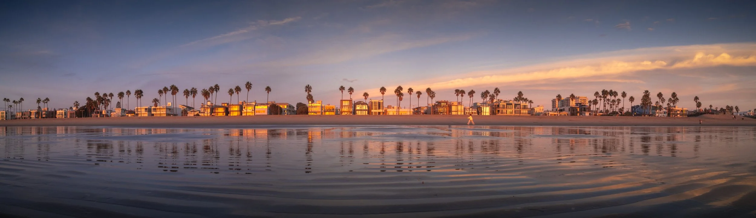 Sunset over a beach with colorful houses lining the shore and palm trees in the background, reflected in the calm water.