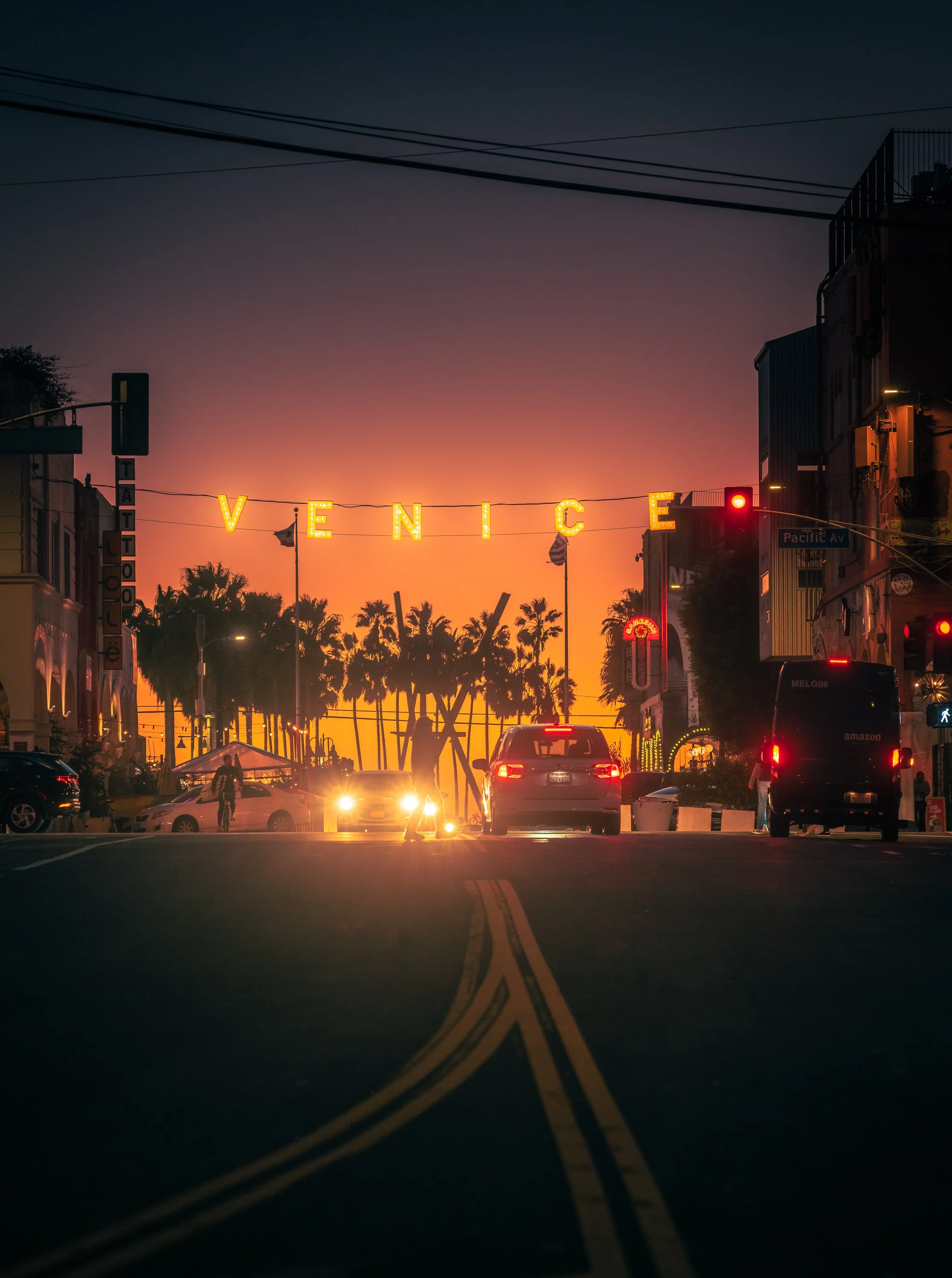 City street at sunset with illuminated sign spelling 'VENICE', palm trees in the background, cars on the road, and traffic lights.