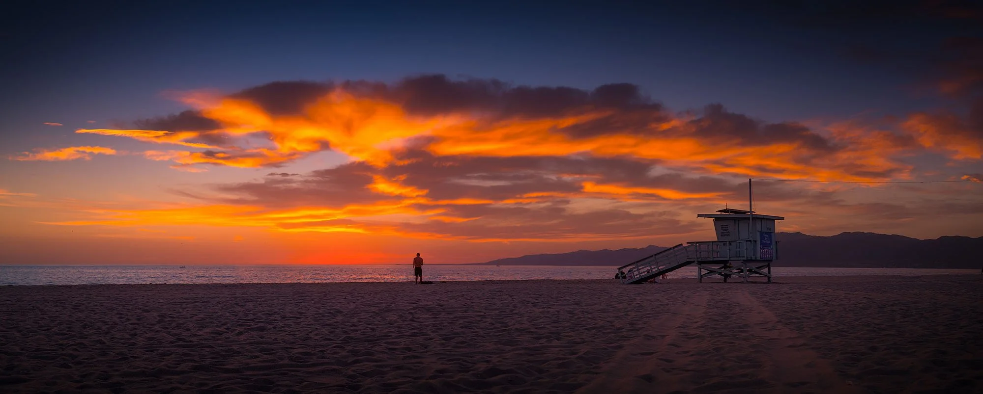 A person standing on the beach watching the sunset with orange and yellow clouds in the sky, a lifeguard tower nearby, mountains in the distance.