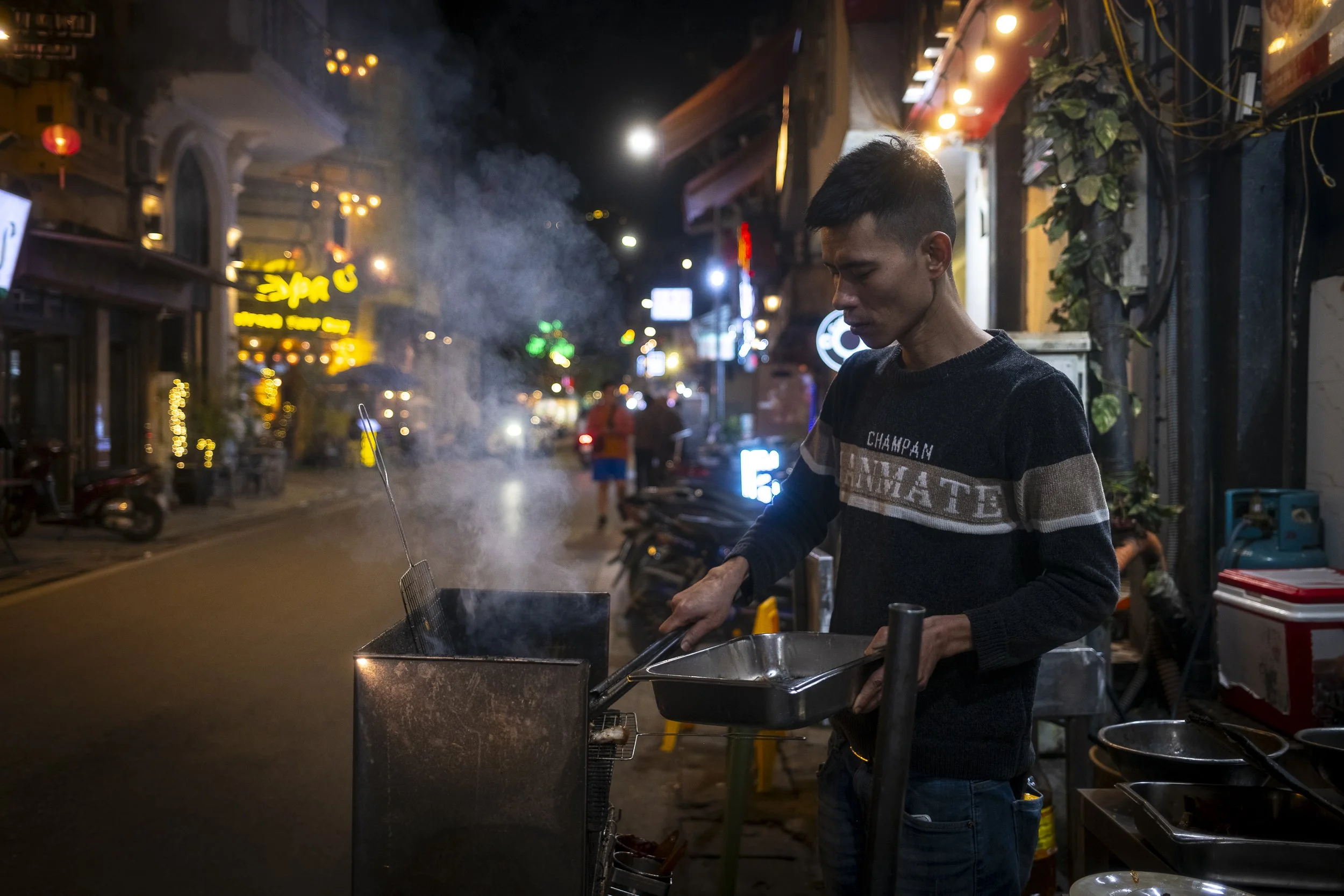 A man cooking food in a street at night, surrounded by neon lights and signs.