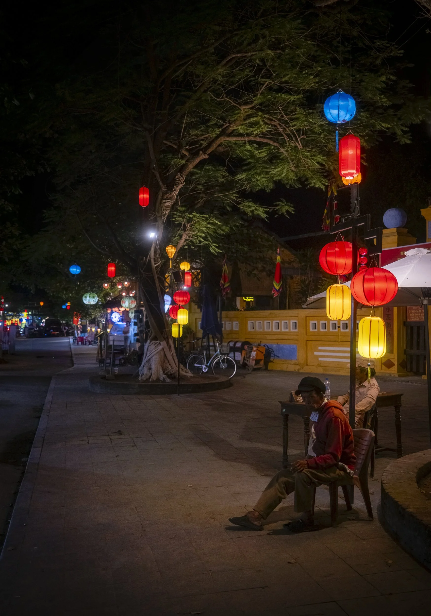 Night scene on a sidewalk with colorful hanging lanterns illuminated in red, yellow, blue, and orange. A man in a red hoodie and cap is sitting on a bench under the lanterns. There are bicycles, trees, and a street with lights and buildings in the ba