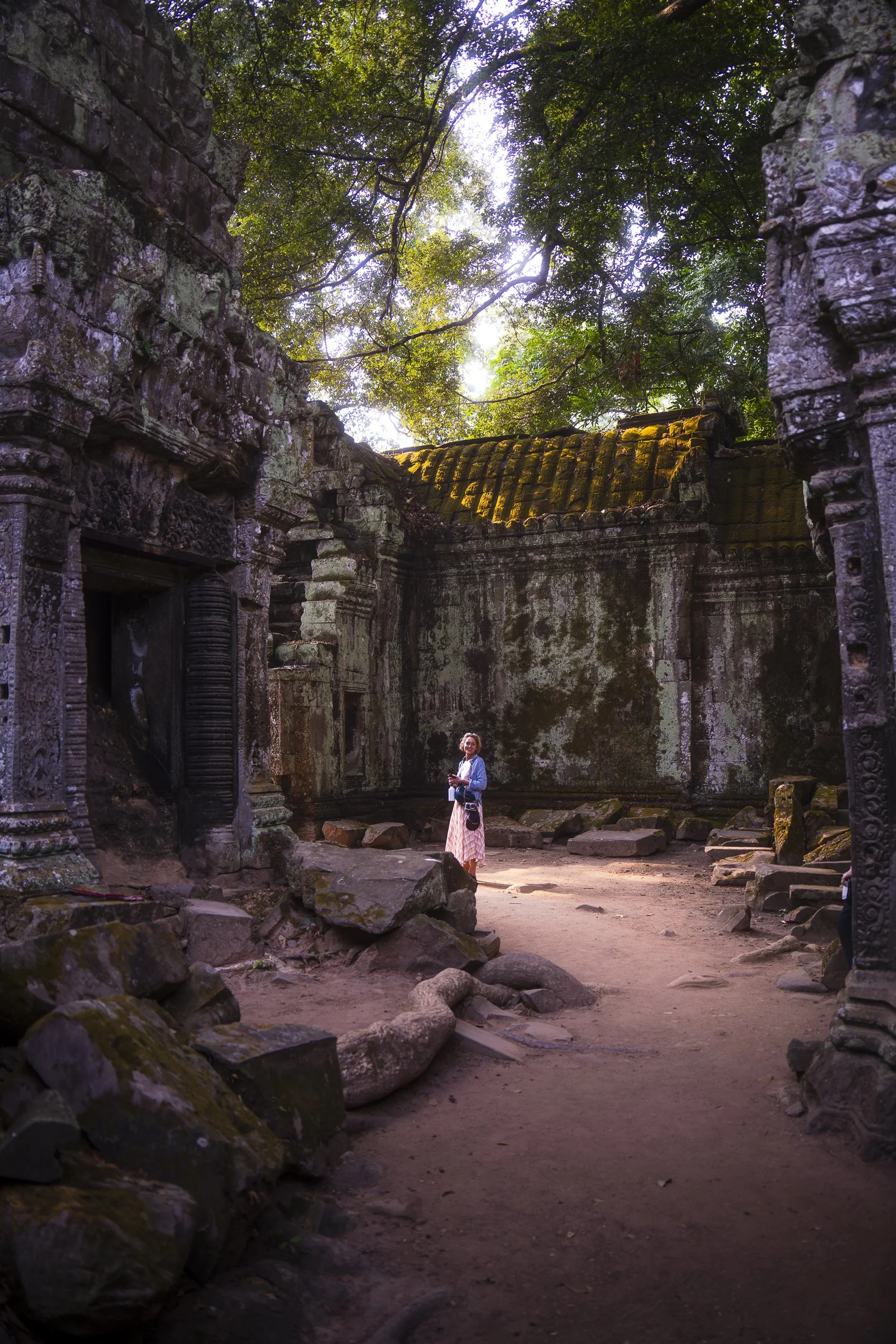 A woman stands inside ancient stone ruins surrounded by moss-covered walls and large rocks, with sunlight filtering through trees overhead.