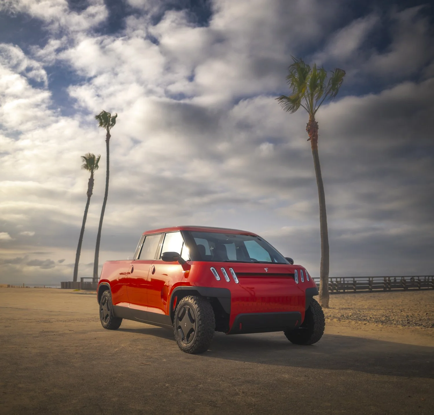 A red futuristic compact vehicle parked on a dirt area at the beach with a backdrop of palm trees, cloudy sky, and a beach fence.