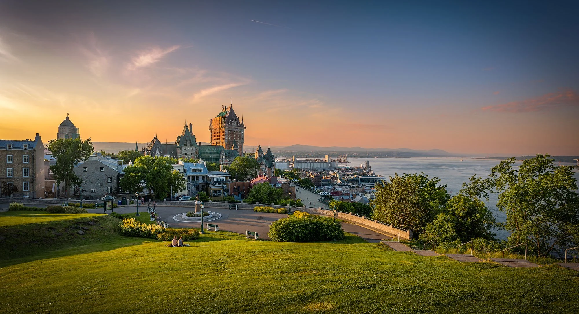 Scenic view of a park overlooking Quebec City with historic architecture, water, and mountains in the background during sunset.