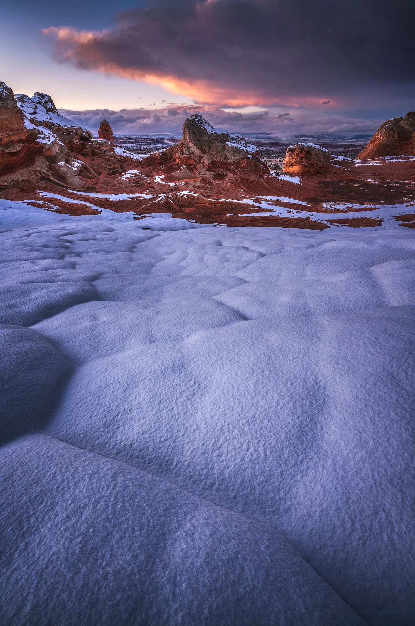 Snow-covered desert landscape with red rock formations and dark cloudy sky during sunset