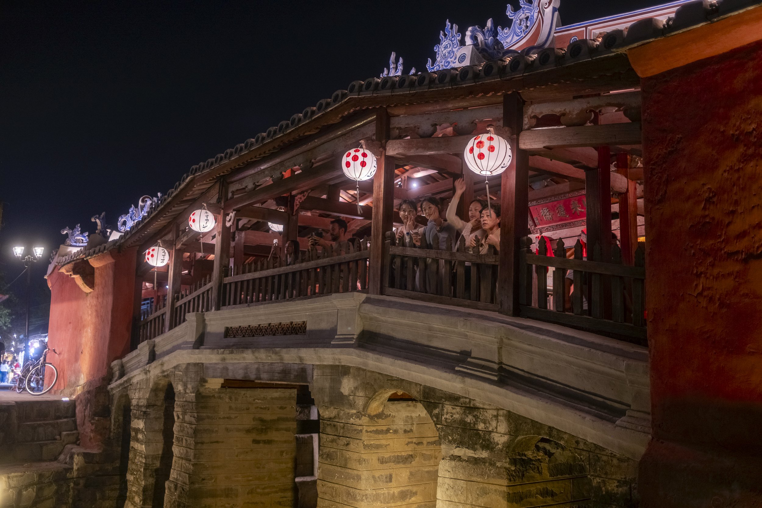 People gathering on a traditional Asian wooden bridge decorated with red and white lanterns at night, with a red wall and ornate roof details.