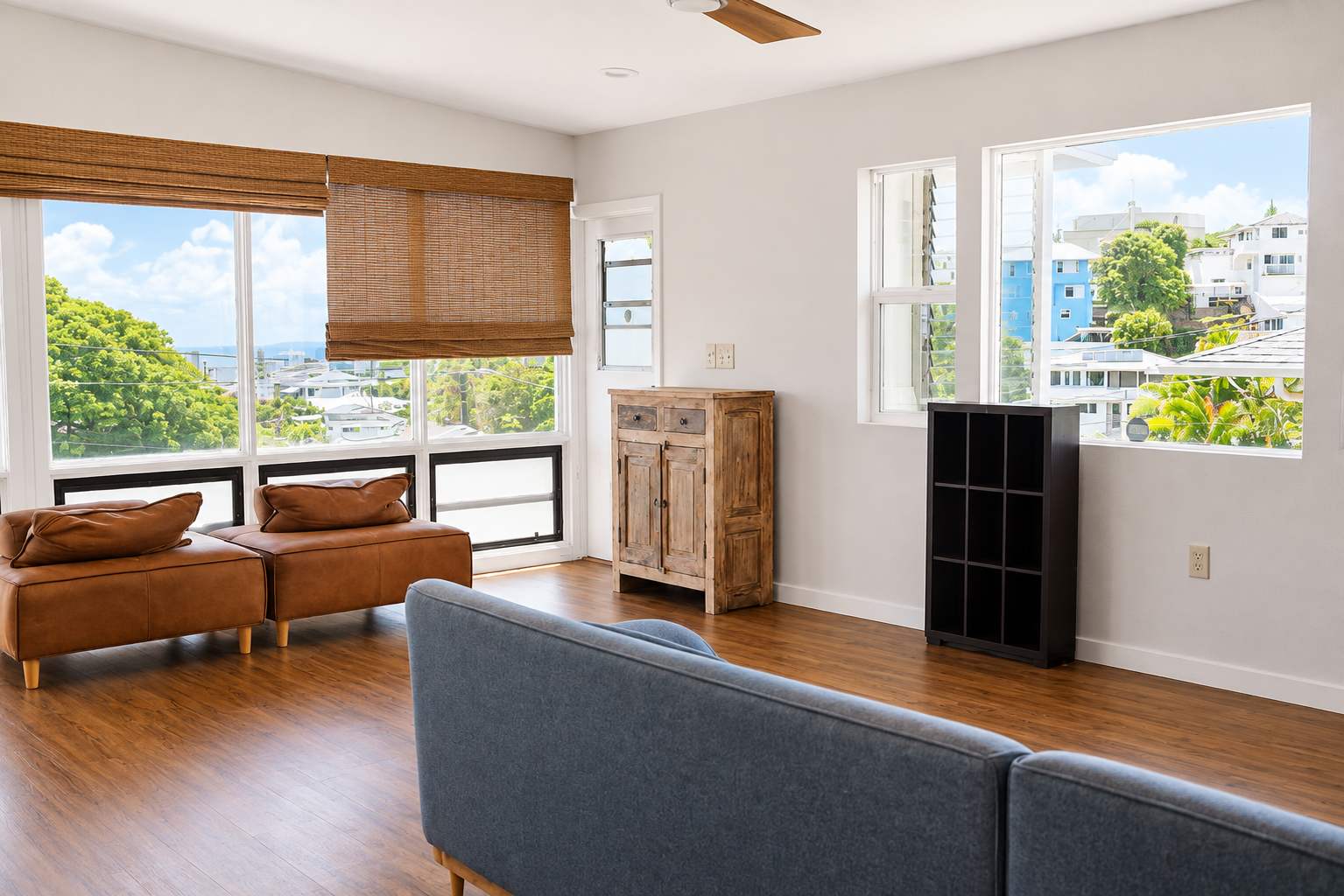 Bright living room with large windows, wooden floors, blue and brown sofas, a wooden cabinet, and a black cubby shelf.