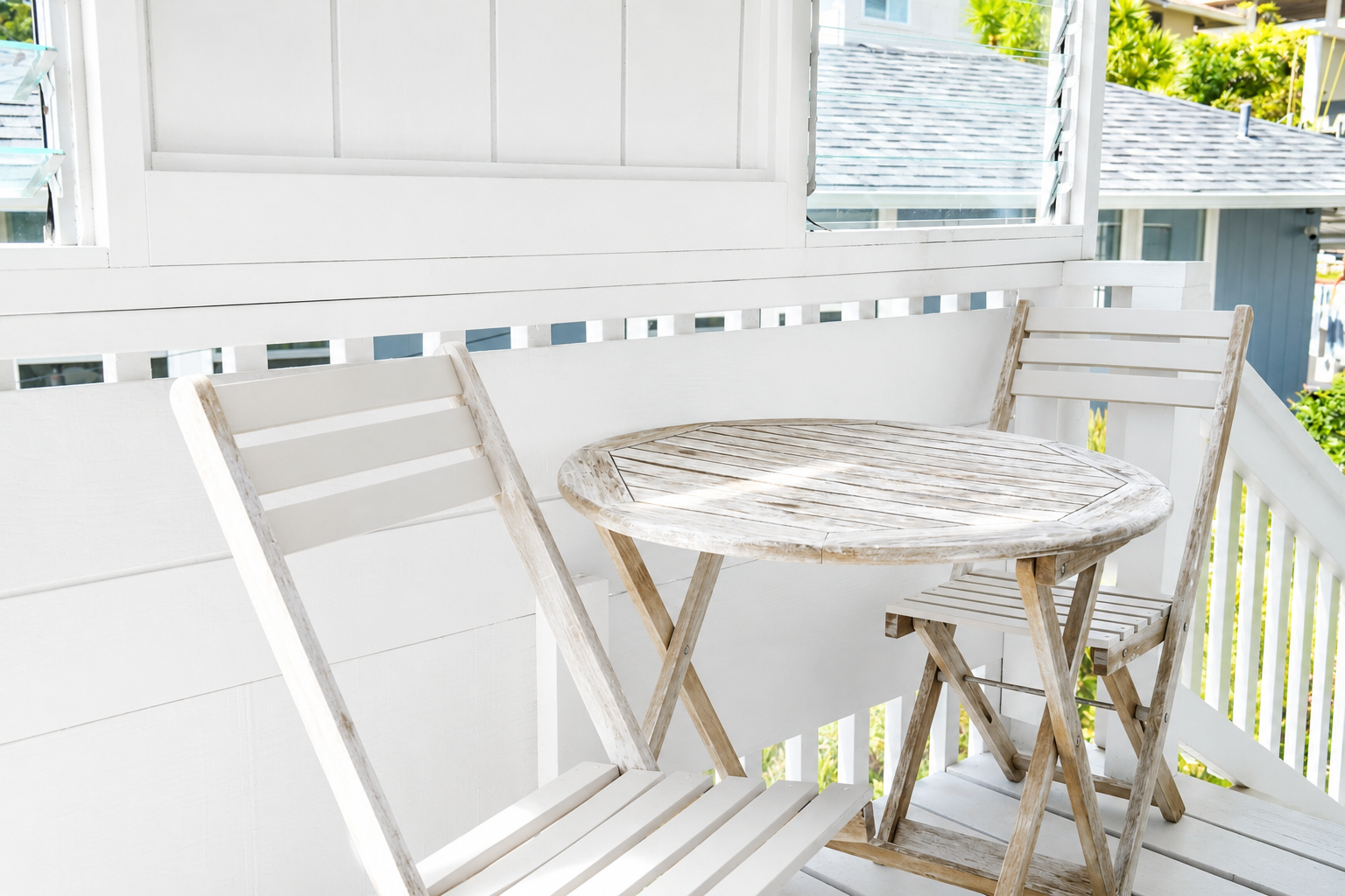 A small outdoor balcony with a round wooden table and two matching wooden chairs, all with a weathered look. The balcony has white fencing and surrounding greenery in the background.