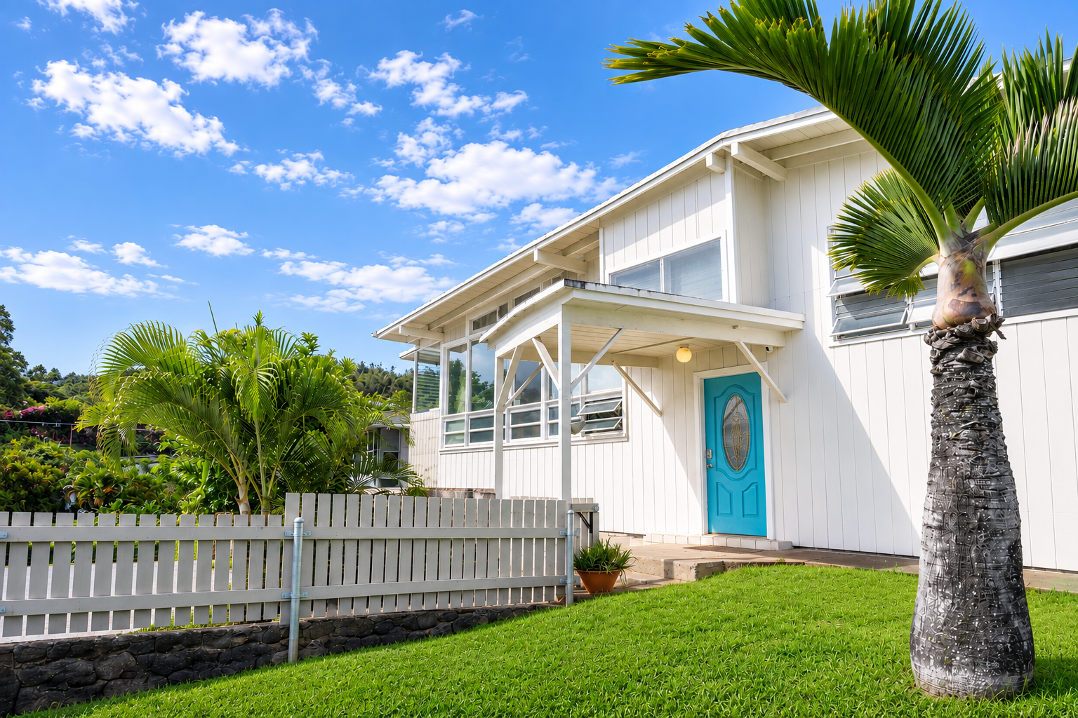 A white two-story house with a bright blue front door, surrounded by lush green grass and tropical plants, including palm trees. The house has large windows and a small porch area with a white railing. The sky is clear with some scattered clouds.