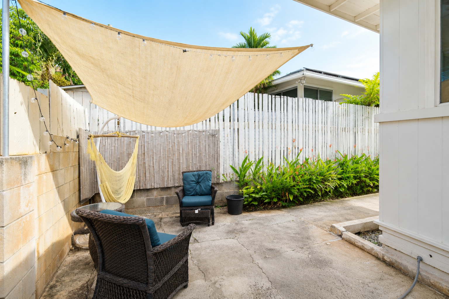 Hawaii Real Estate Photography: Cozy backyard patio with two wicker chairs and blue cushions, a hanging yellow hammock, string lights, lush green plants with orange flowers, and a beige canopy, enclosed by a white wooden fence.