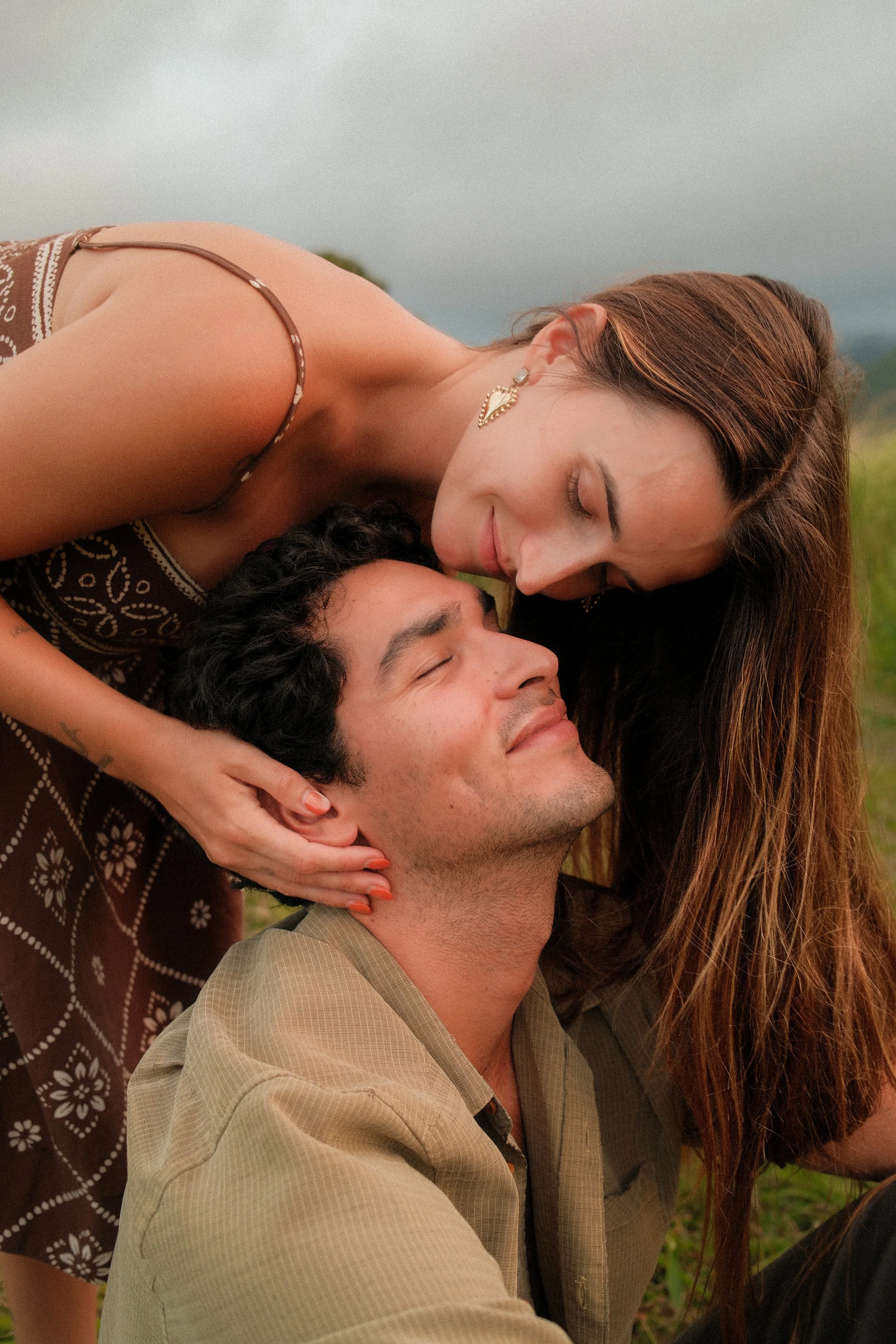 A woman with long brown hair wearing earrings and a patterned dress, leaning down to kiss a man with dark curly hair and a beige shirt, in an outdoor setting with a blurred background.