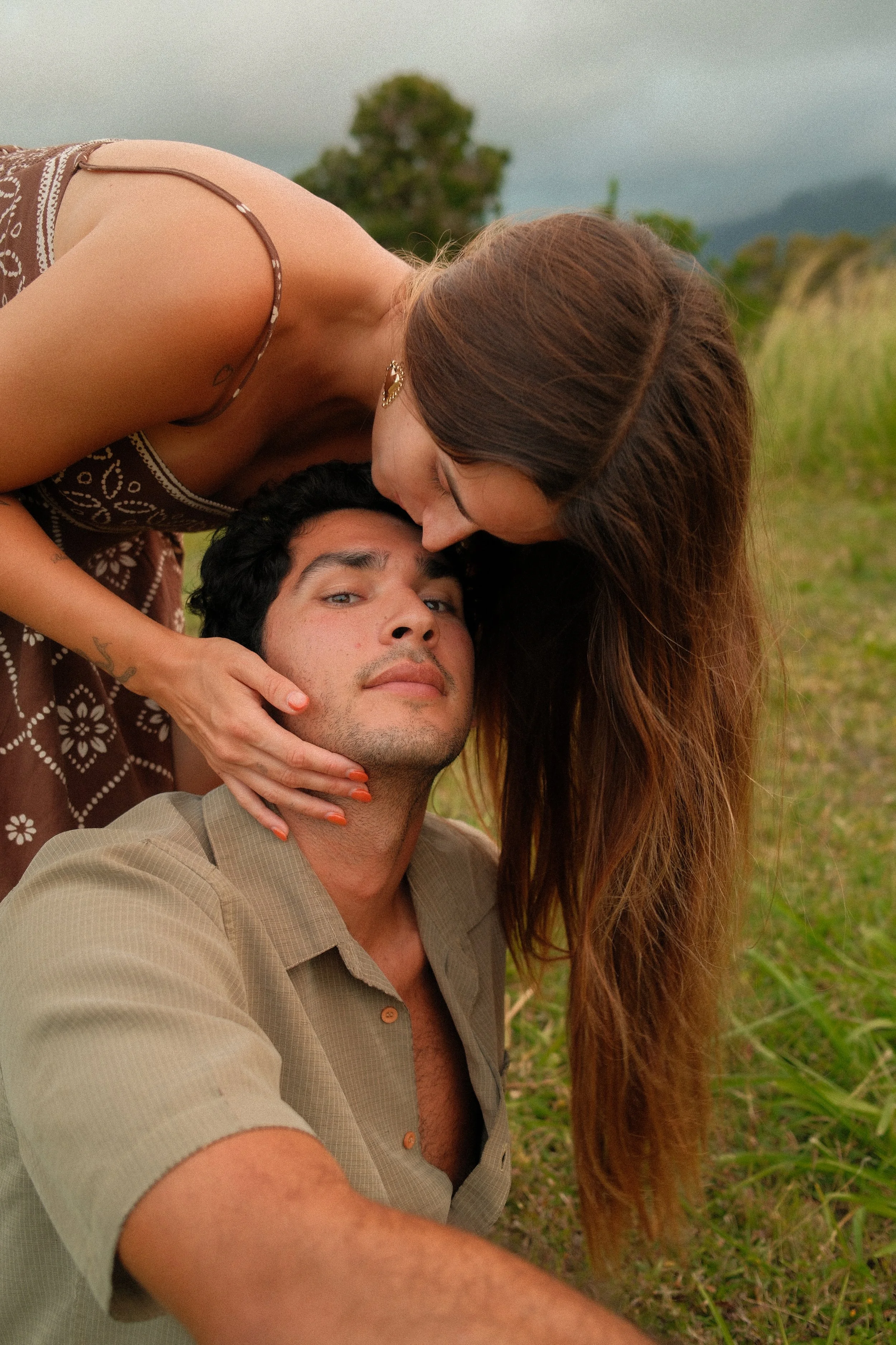 A couple lying together outdoors in a grassy area, with the woman kissing the man on the forehead. The man is looking at the camera and wearing a beige shirt, while the woman has long brown hair and is wearing a patterned dress.