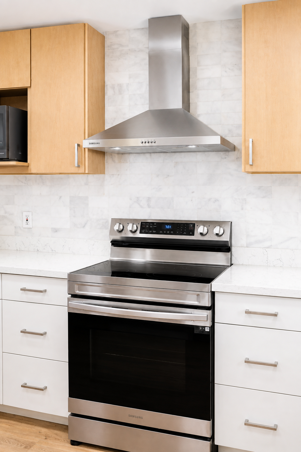 Stainless steel Samsung stove in a modern kitchen with white countertops, white drawers, light wood cabinets, a tiled white backsplash, and a metal range hood.